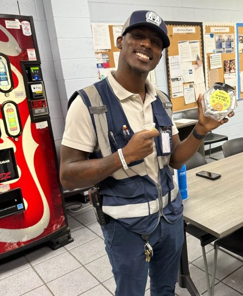 A smiling man in uniform holding a small jar with a yellow sticker that says 'Sunshine in a Jar.' He is standing indoors near a vending machine and a bulletin board.