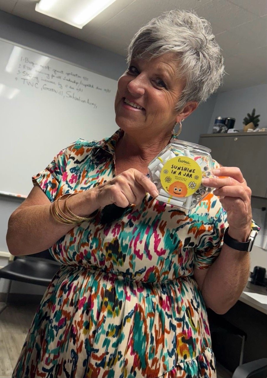 A smiling elderly woman with short gray hair wearing a colorful patterned dress holding a small jar labeled "Sunshine in a Jar" with a cartoon sun and smiling face, in an office setting.