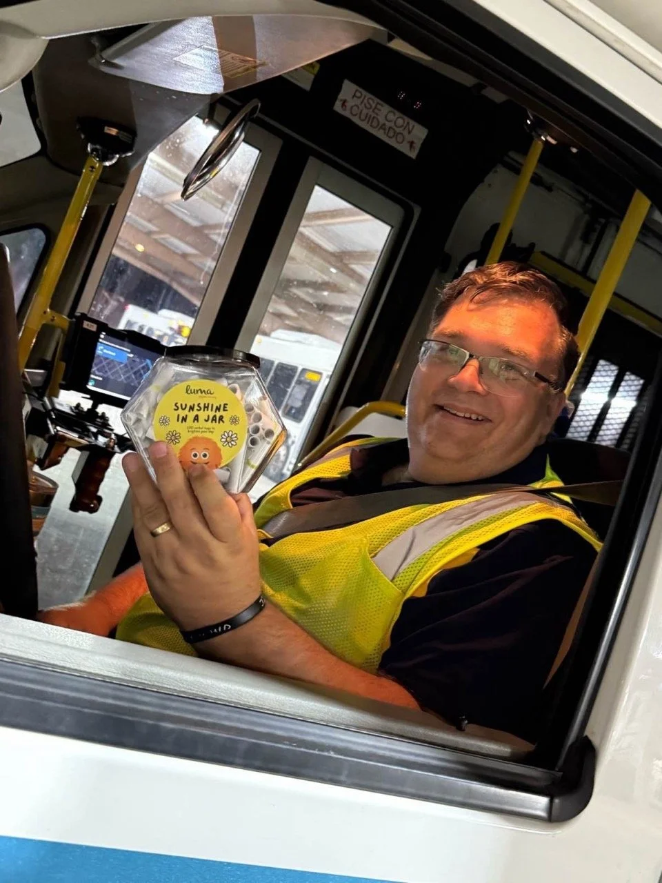 A smiling man in a yellow safety vest and glasses sitting inside a bus, holding a small, clear container labeled 'Sunshine in a Jar' with a cartoon face and yellow flowers.