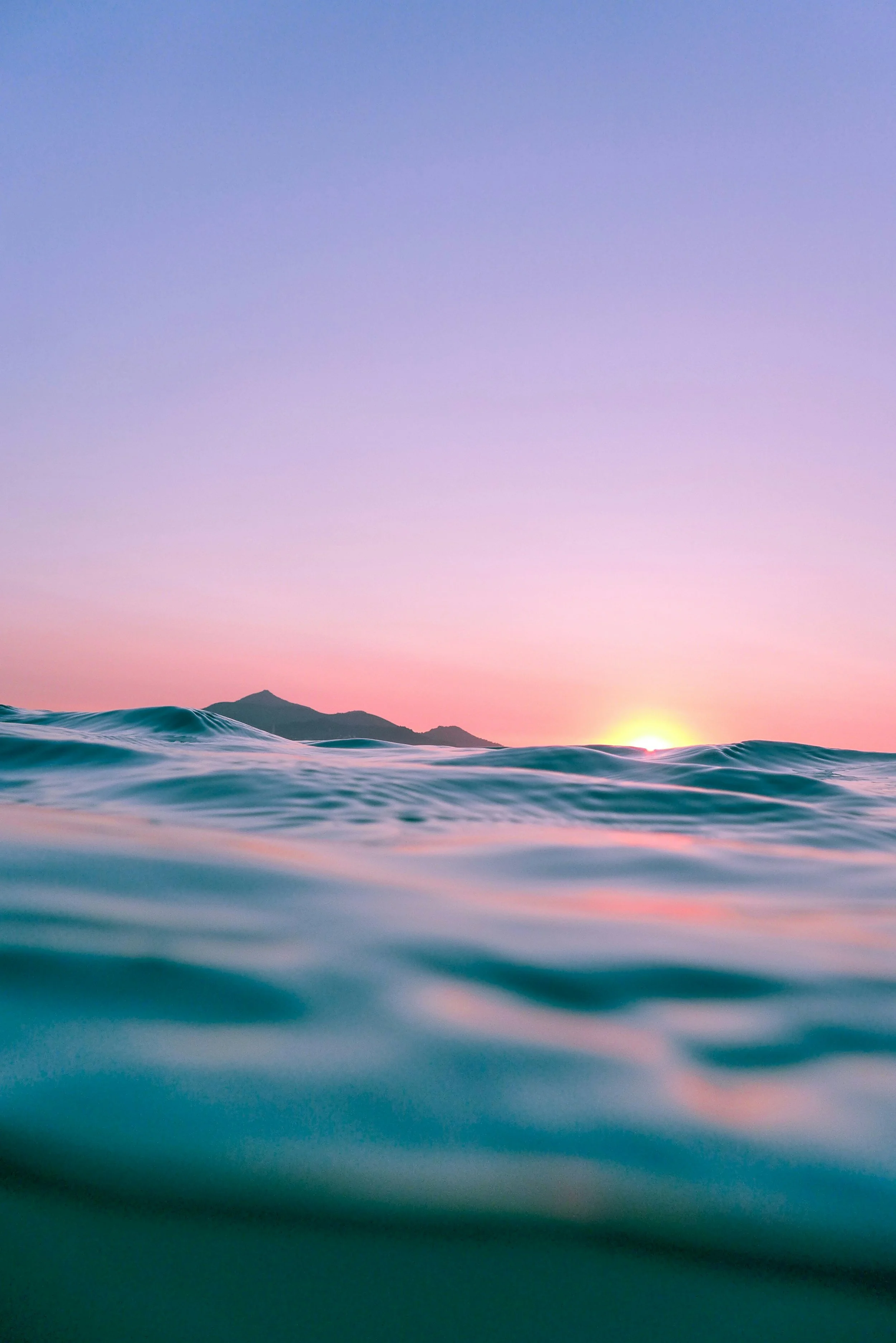 Sunset over the ocean with gentle waves and a silhouette of distant mountains in the background.