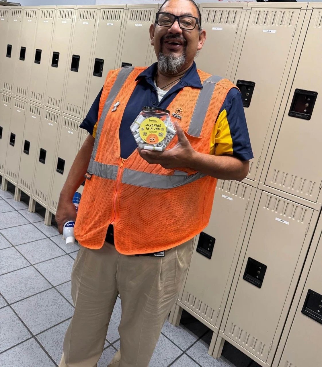 A man wearing glasses and an orange safety vest smiling and holding a jar labeled 'Sunshine in a Jar' in a locker room.