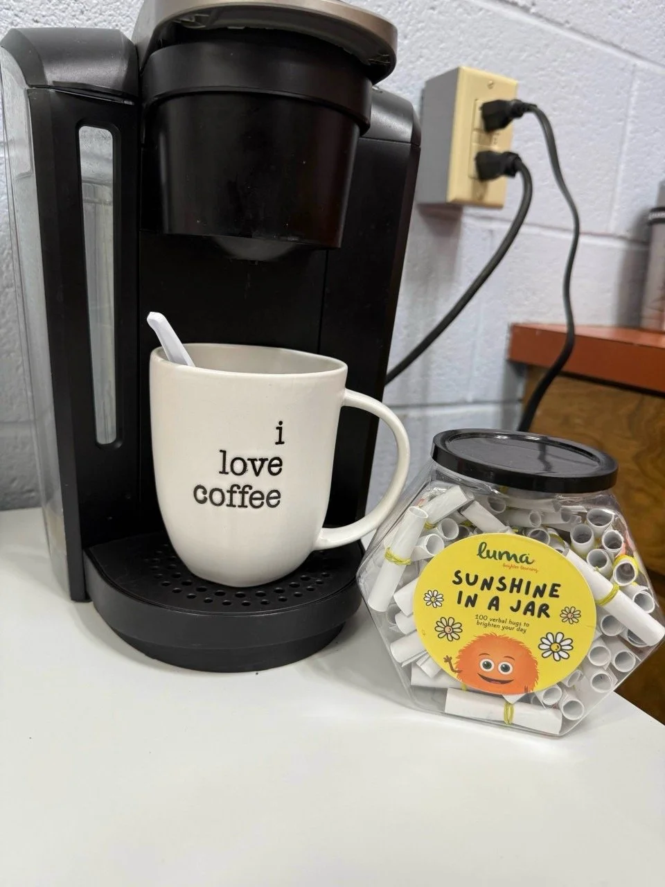 Black coffee maker with a white mug reading 'I love coffee' placed underneath the spout, a container labeled 'Sunshine in a Jar' filled with paper straws, and a plugged-in wall outlet on a white countertop against a cinder block wall.