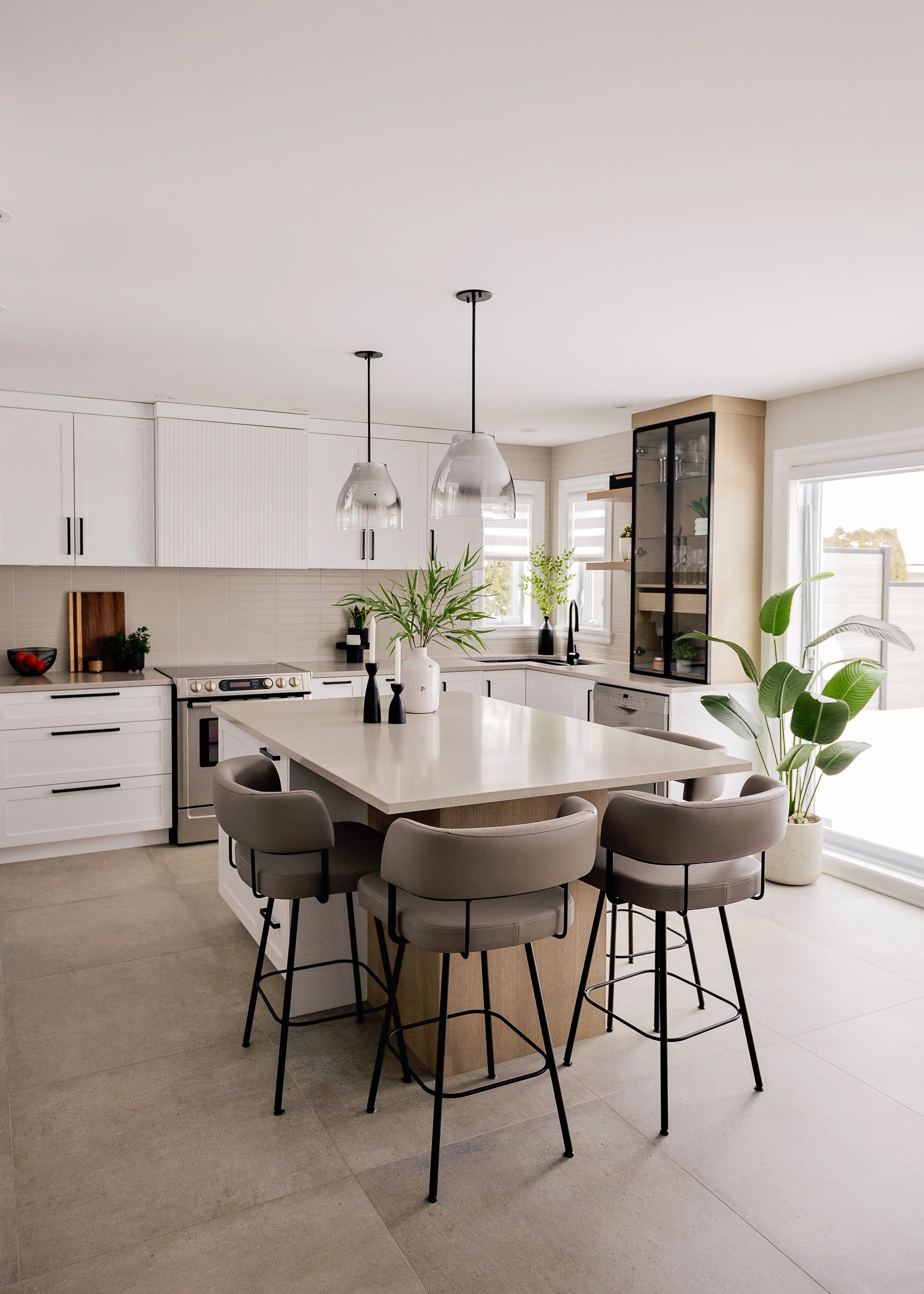 Cuisine moderne avec un plan de travail blanc, des chaises grises autour d'une table centrale, des armoires blanches, des plantes, deux lampes suspendues et une grande fenêtre apportant de la lumière naturelle.