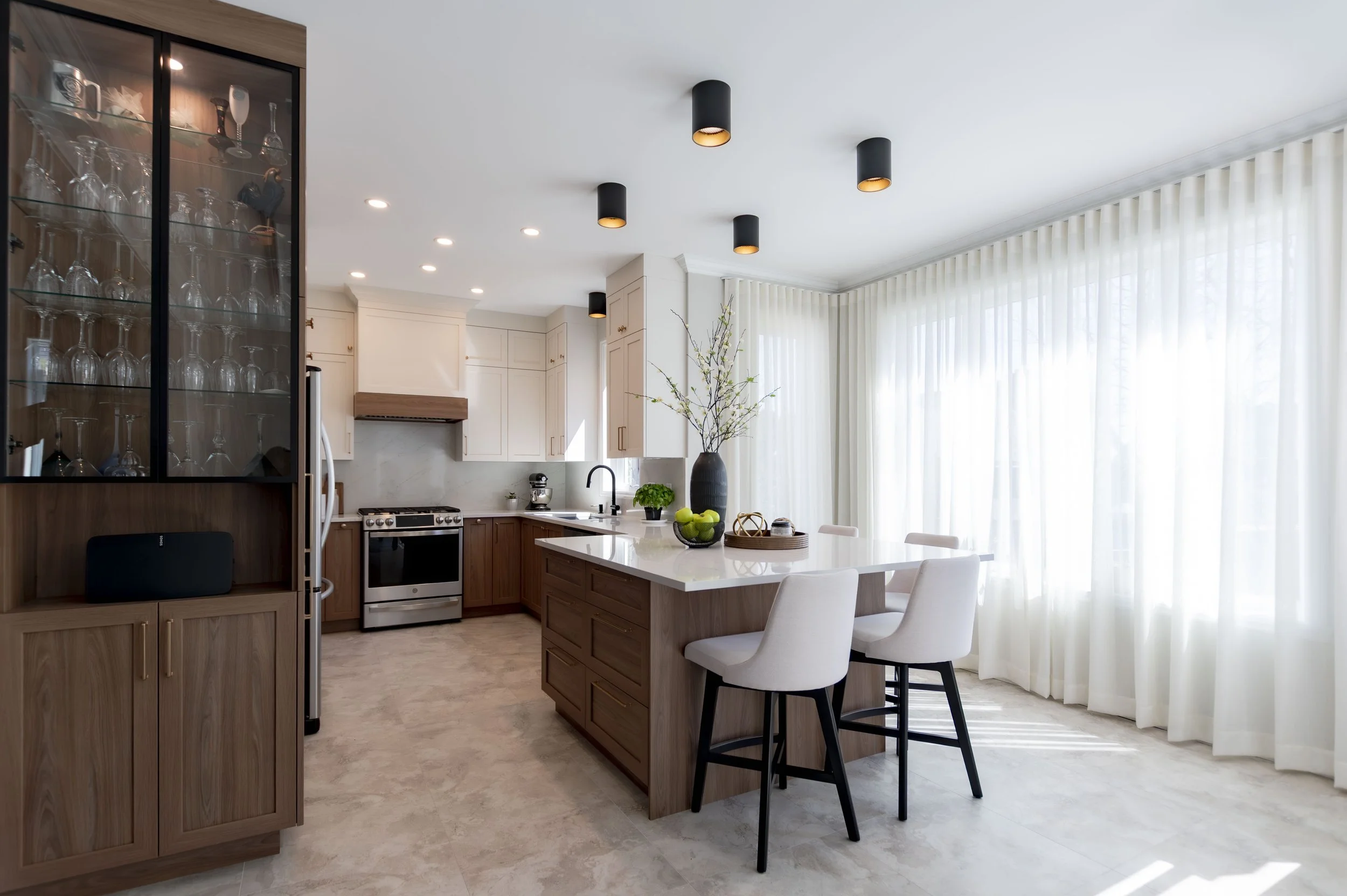 Cuisine moderne avec armoires en bois clair, plan de travail en quartz blanc, éclairage encastré, un grand îlot central avec quatre chaises, grande fenêtre avec rideaux blancs, décor minimaliste avec un vase et des pommes de terre sur l'île.