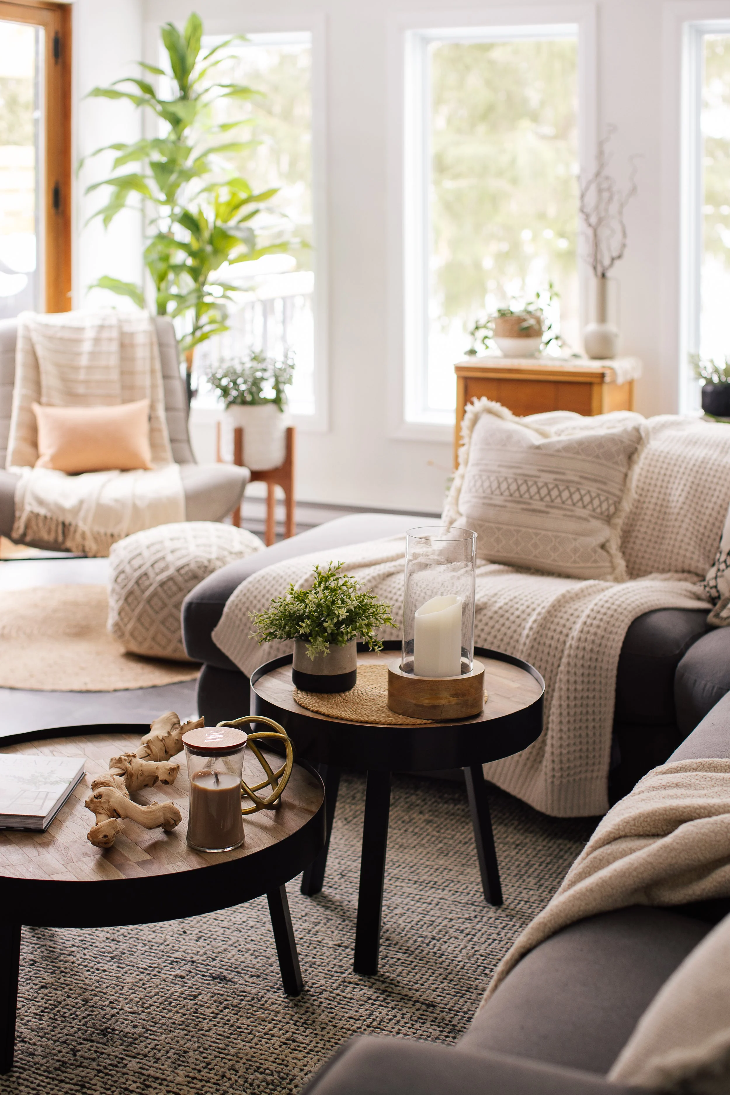 Salon lumineux avec meubles en tissu beige, plantes vertes, tables basses en bois avec bougies, verres, et objets décoratifs, tapis beige, fenêtres larges laissant entrer la lumière naturelle.