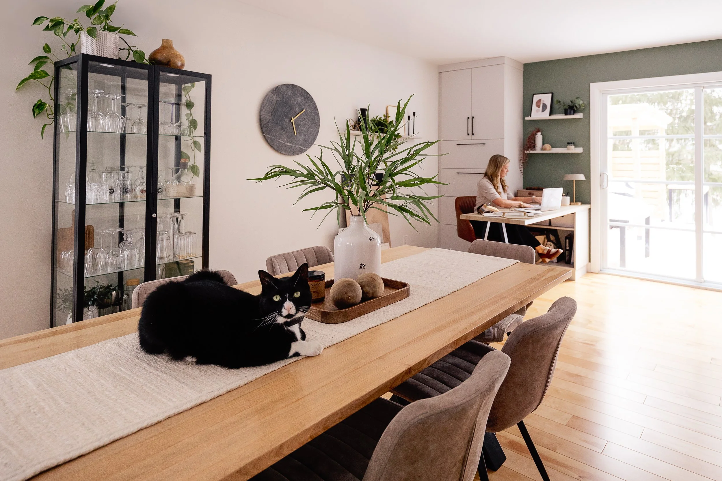 Une table en bois rectangulaire avec une nappe beige. Un chat noir et blanc est allongé sur la table avec des balles de bois dans un plateau en bois. En arrière-plan, une grande plante verte dans un vase blanc, un mur blanc avec une horloge ronde et un placard blanc, un mur vert avec des étagères et une fenêtre en baie vitrée laissant entrer la lumière naturelle. Une femme travaille à un bureau près de la fenêtre.
