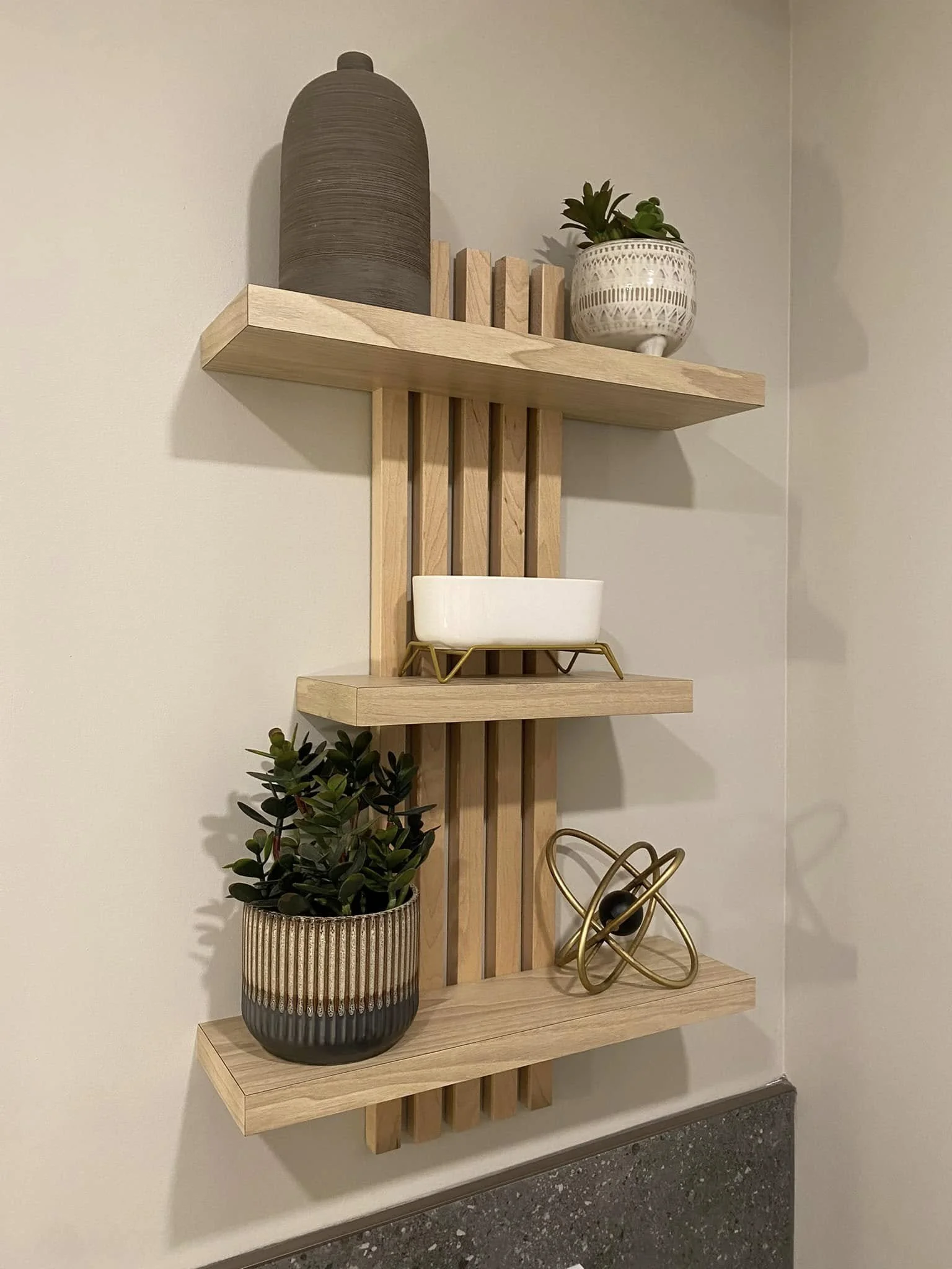 Une étagère en bois munie de plusieurs décorations, dont un vase noir avec des plantes vertes, un pot blanc avec une plante succulente, une sculpture en métal doré, et un pot en osier avec une plante verte.