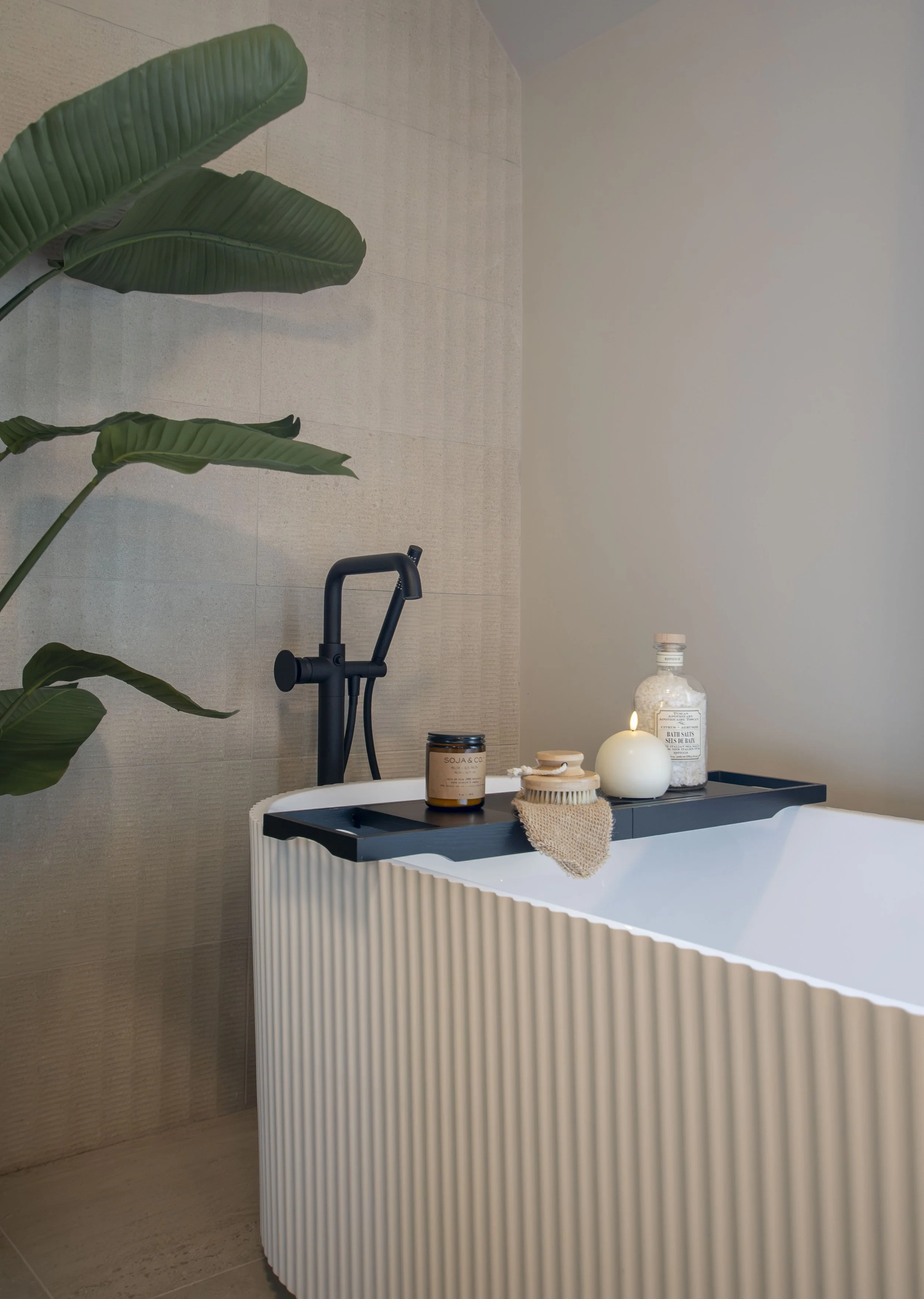 Une salle de bain moderne avec une baignoire blanche à surface ondulée, un mur beige derrière, un grand plant vert à gauche, et une étagère noire à droite avec des objets décoratifs comme une bougie, une bouteille de bain, et un pot en terre cuite.