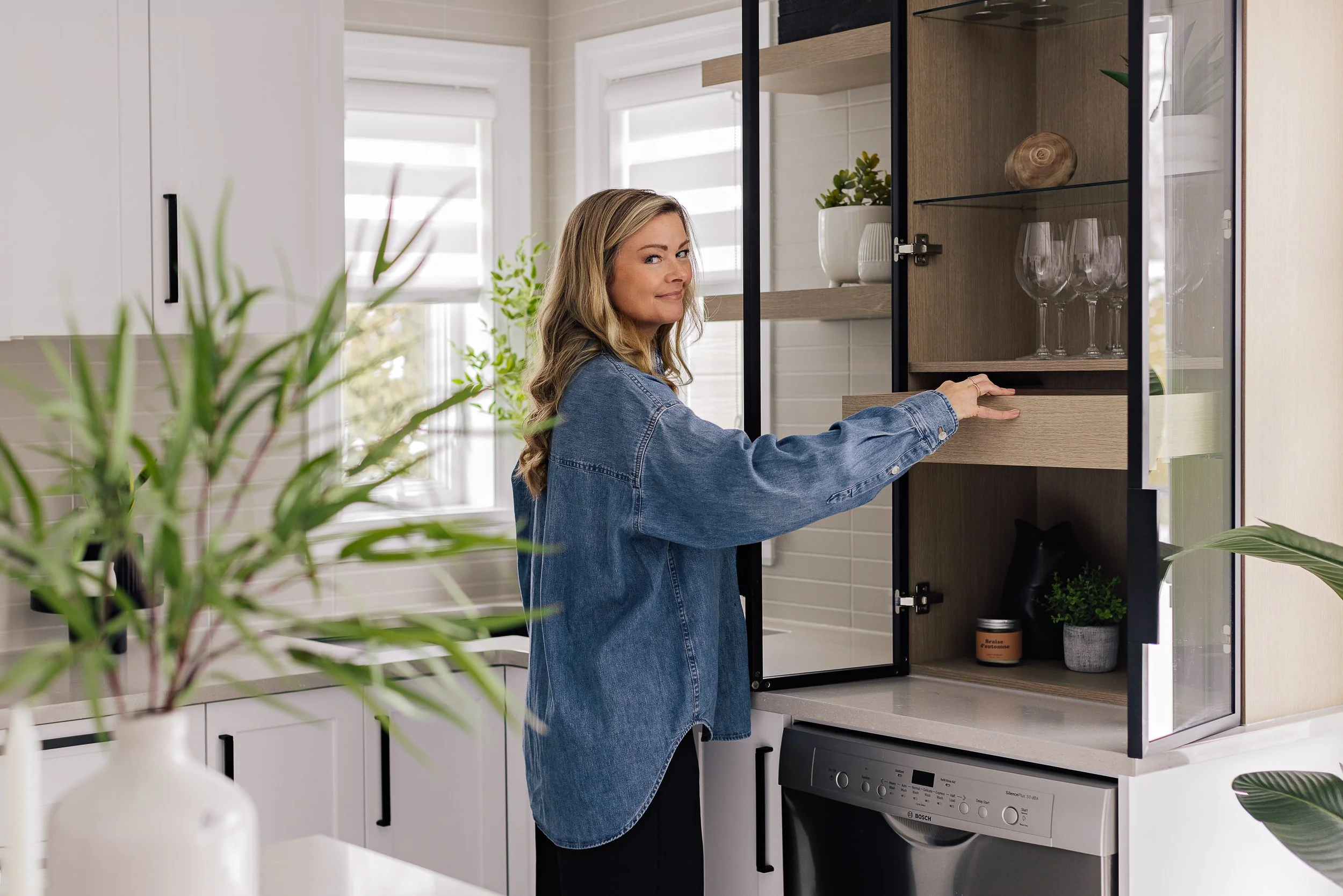 Chantal Rouiller - designer d'intérieur indépendante à Saint-Jean-sur-Richelieu, ouvre un placard de cuisine ou se trouve de la vaisselle, des plantes et des éléments décoratifs.