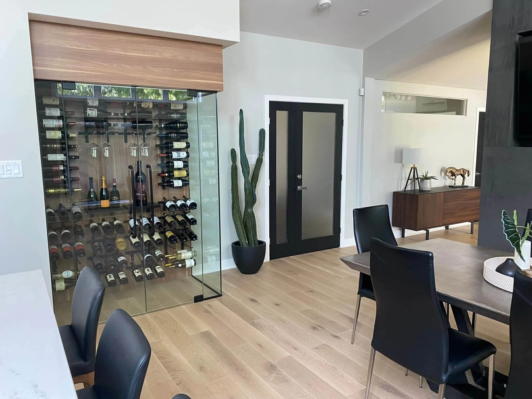 Une pièce moderne avec une cave à vin en verre, un cactus, une porte noire, une œuvre d'art, un tapis rond, une table et des chaises.