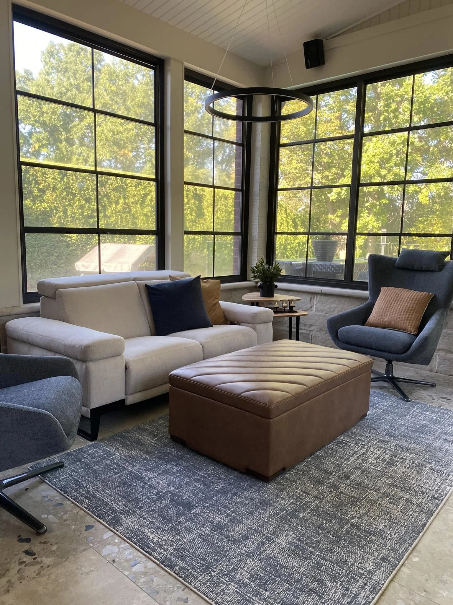 Salon avec un canapé blanc, un fauteuil gris, un ottoman marron, une table basse ronde, un tapis gris et une grande fenêtre laissant entrer la lumière naturelle, avec des arbres visibles à l'extérieur.