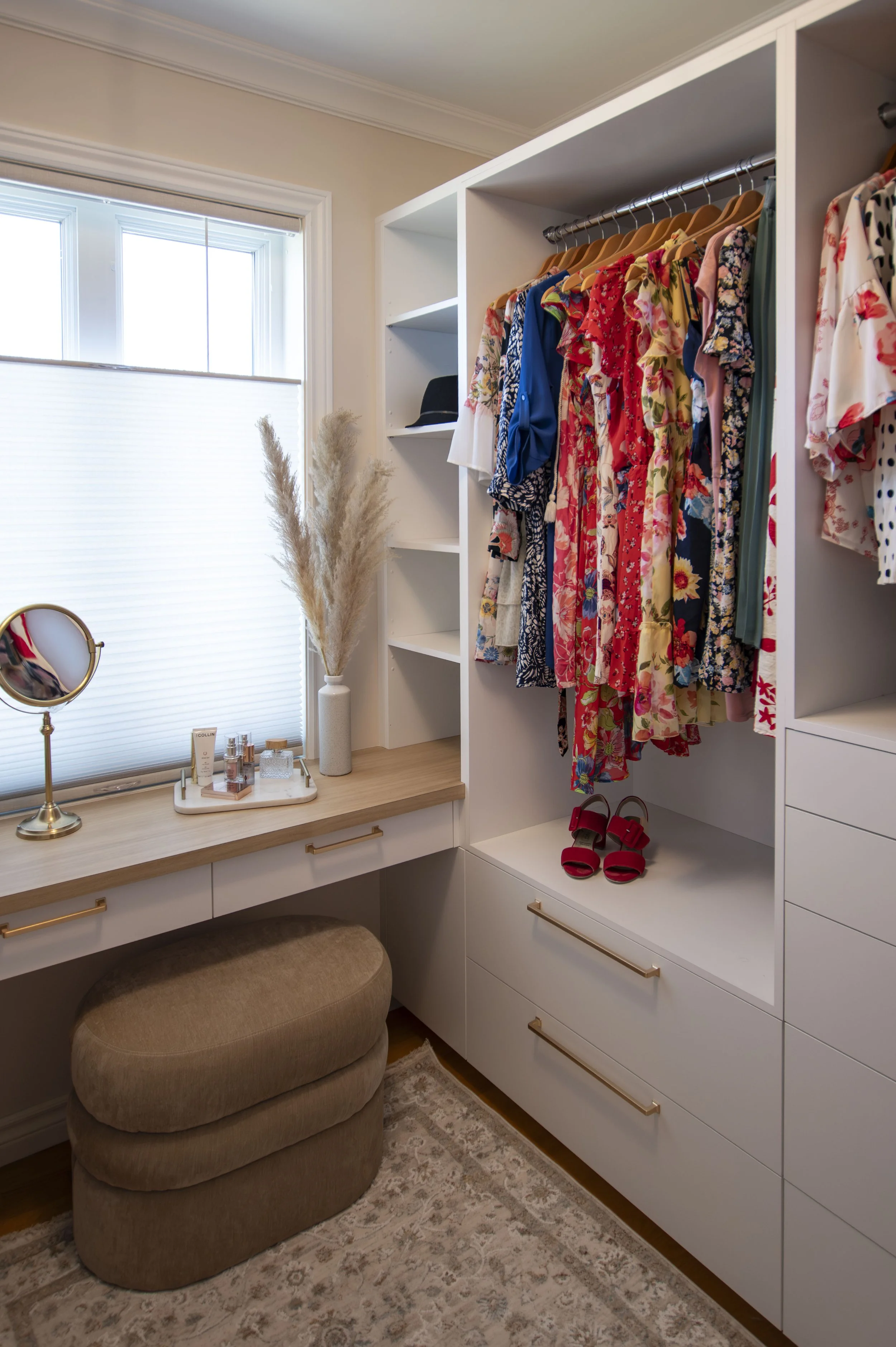 Une chambre avec un placard blanc contenant des vêtements colorés suspendus, une paire de sandales rouges sur une étagère, un bureau en bois clair avec un miroir, une plante en paille, et un fauteuil beige. La pièce est bien éclairée avec une grande fenêtre laissant entrer la lumière naturelle.