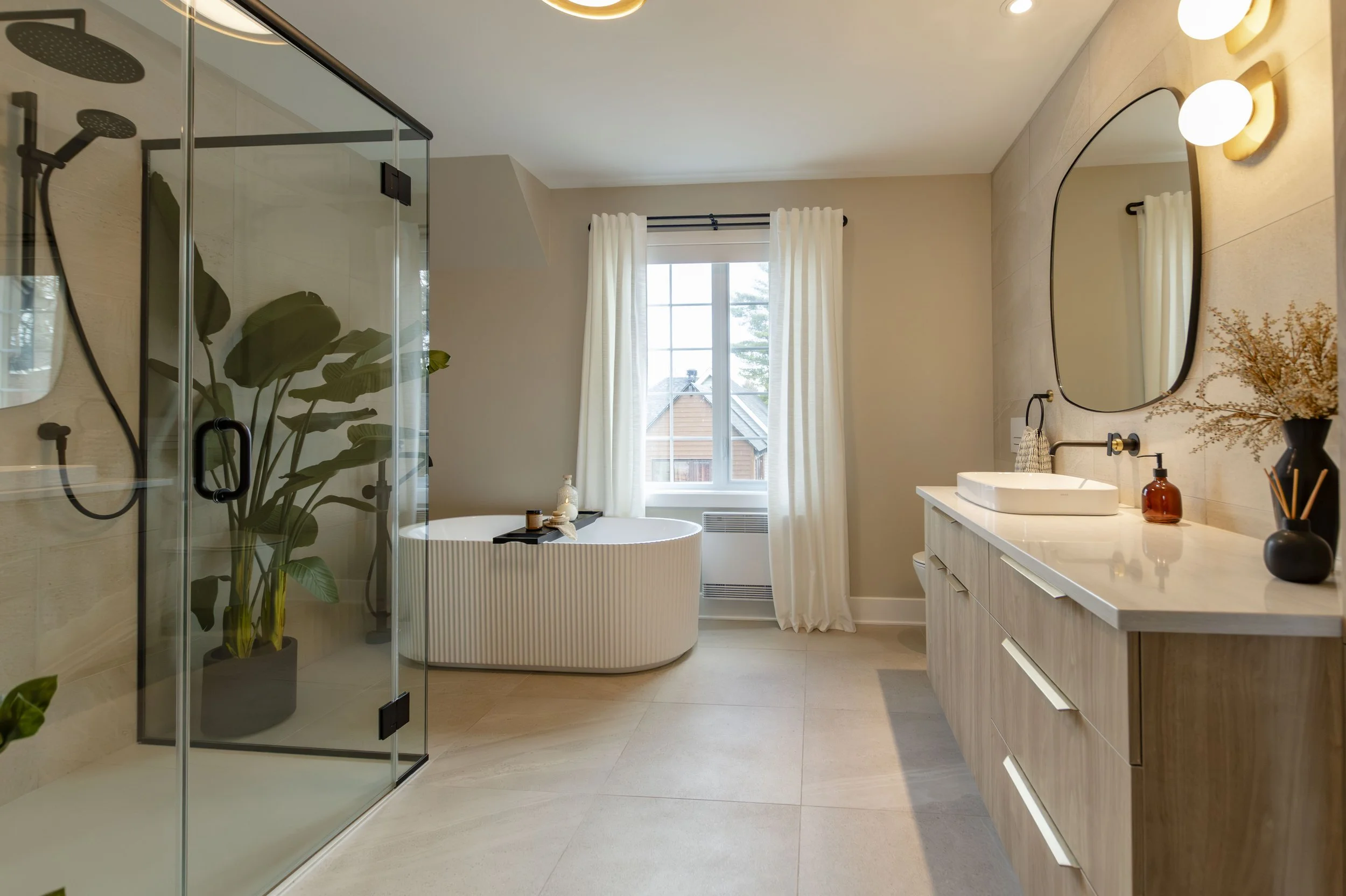 Salle de bain moderne avec douche en verre, baignoire sur pattes, grand miroir, lavabo, et décorations végétales et lumineuses.