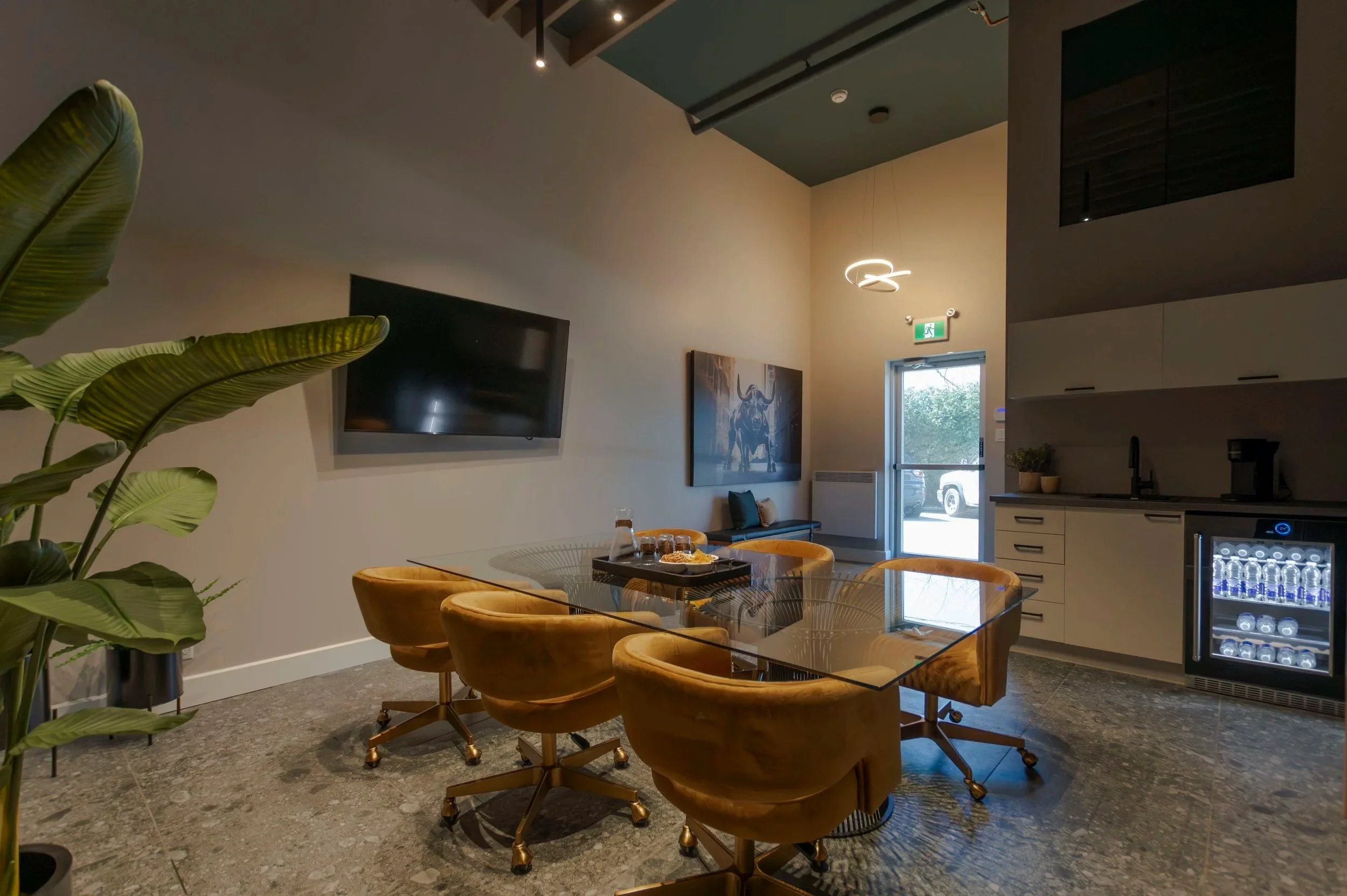 Intérieur d'une salle de réunion ou de détente avec un cercle de fauteuils en velours orange autour d'une table en verre, une télévision murale, une peinture représentant un taureau, et une kitchenette avec un réfrigérateur rempli de bouteilles d'eau.