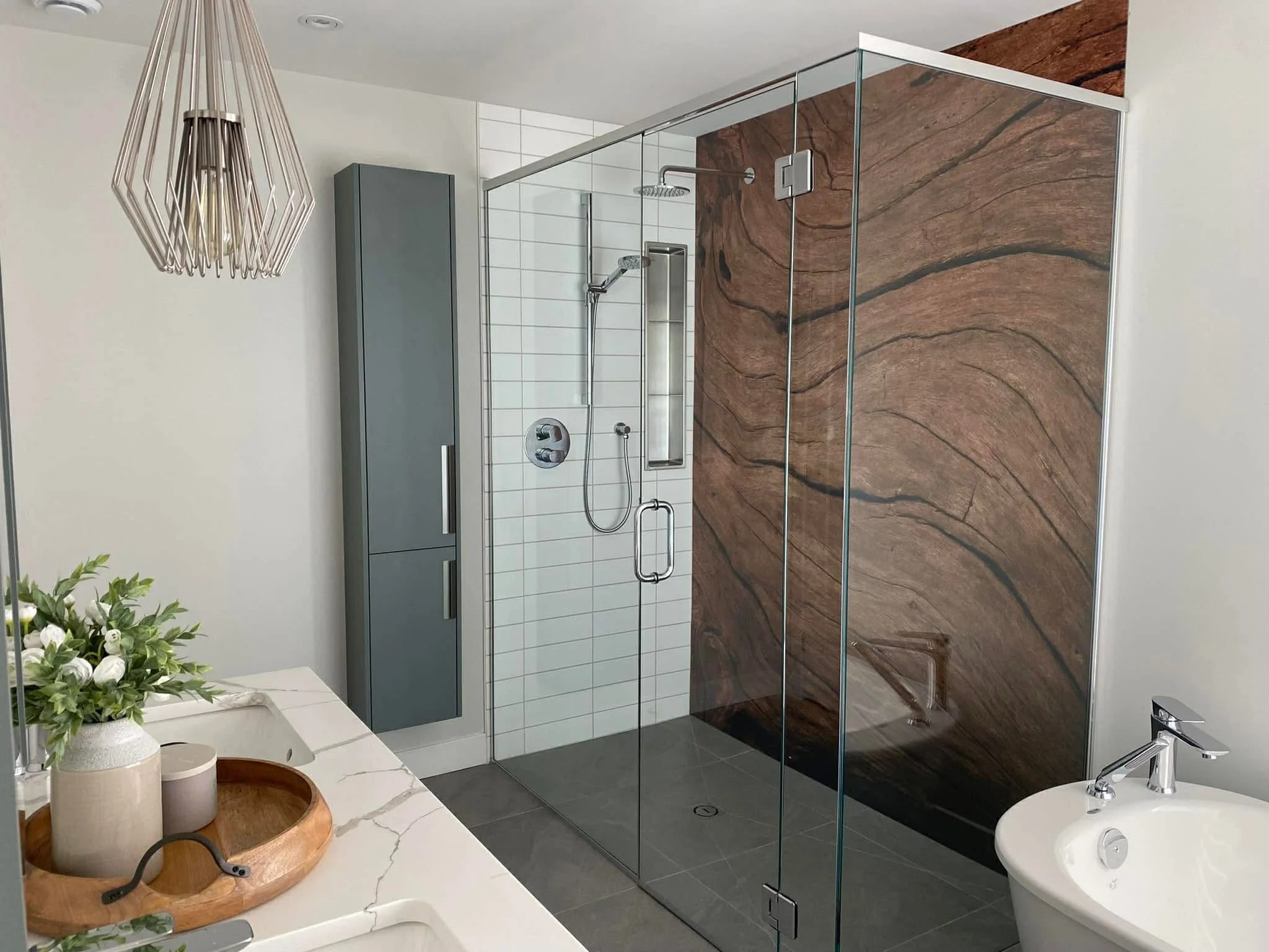 Une salle de bain moderne avec une douche à l'italienne avec parois en verre, un mur en pierre marron, un meuble gris, un lavabo rond blanc avec un robinet, des fleurs dans un vase blanc sur une surface en marbre, et un éclairage suspendu design.