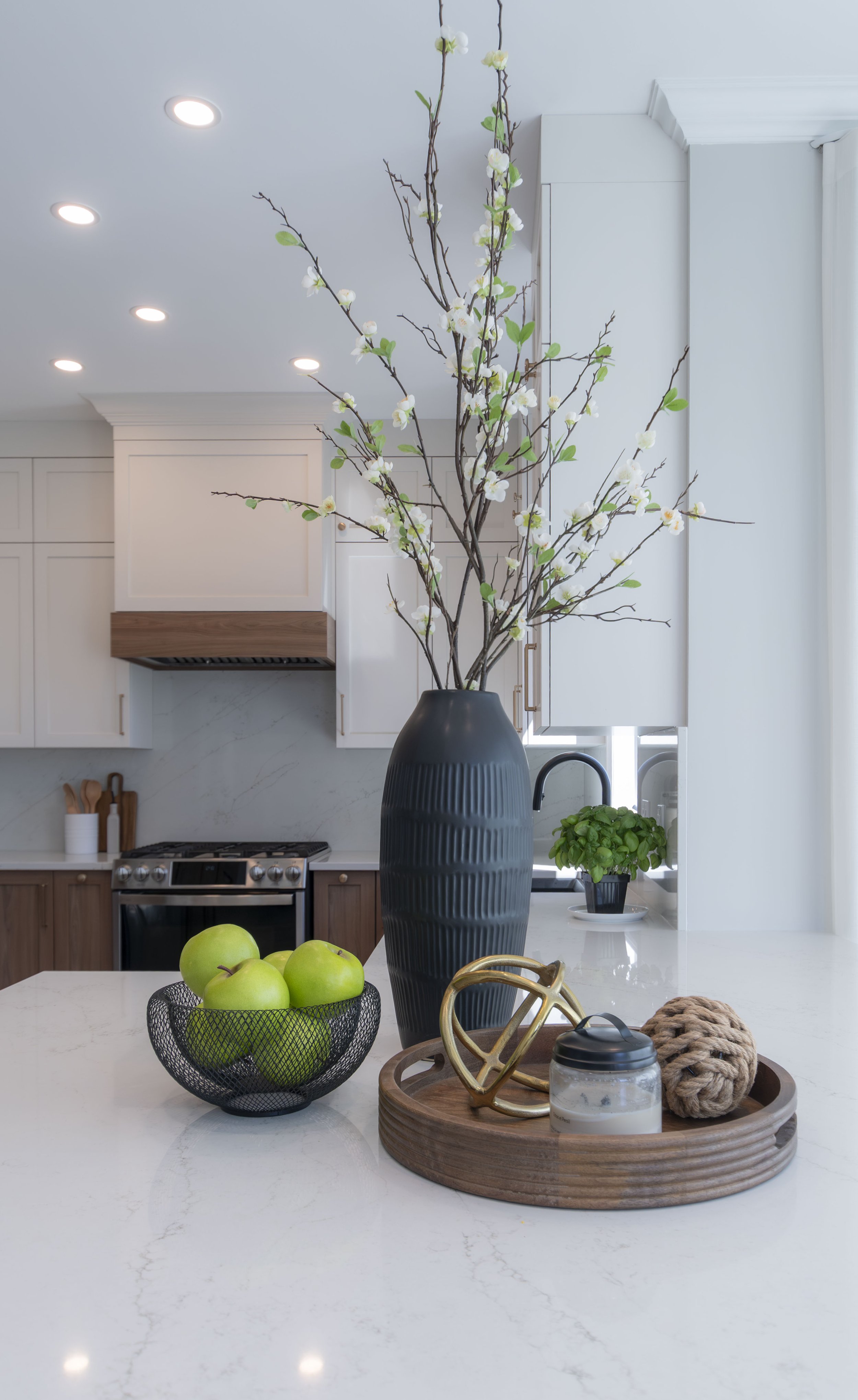 Vase noir avec branches florales blanches et vertes sur une table de cuisine blanche. Autour, une coupelle avec pommes vertes, un pot de basilic, et un plateau en bois avec un autour décoratif, une bougie, et une pelote de corde. En arrière-plan, cuisine blanche avec appareils en acier inoxydable et lumière encastrée.
