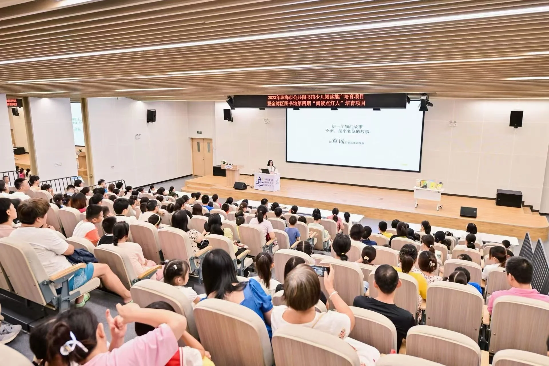 Children and adults seated in an auditorium watching a presentation on a large screen.