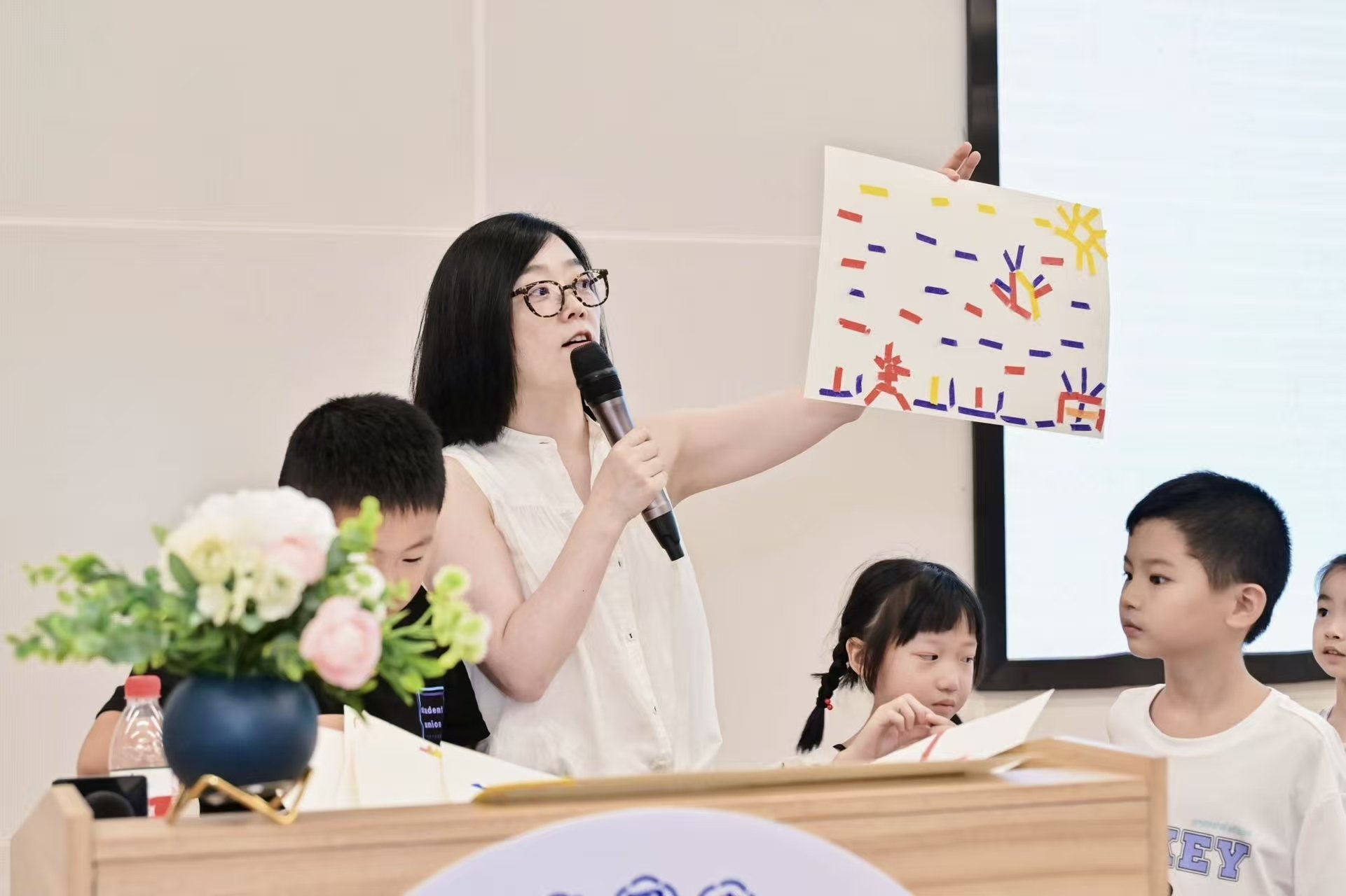 A woman with glasses holding a microphone and a colorful drawing, speaking to children at a classroom or event. Children are looking at her, with one girl looking at her with interest, and a boy looking at her attentively. There is a vase with flowers on the table in front of her.