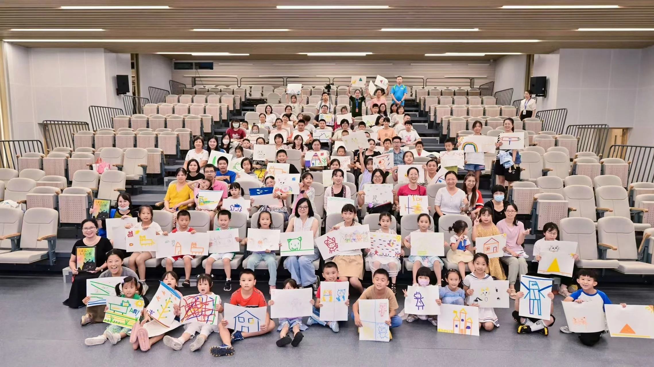 A large group of children and adults sitting and standing in an auditorium, holding up drawings and artwork, with some smiling and looking at the camera.