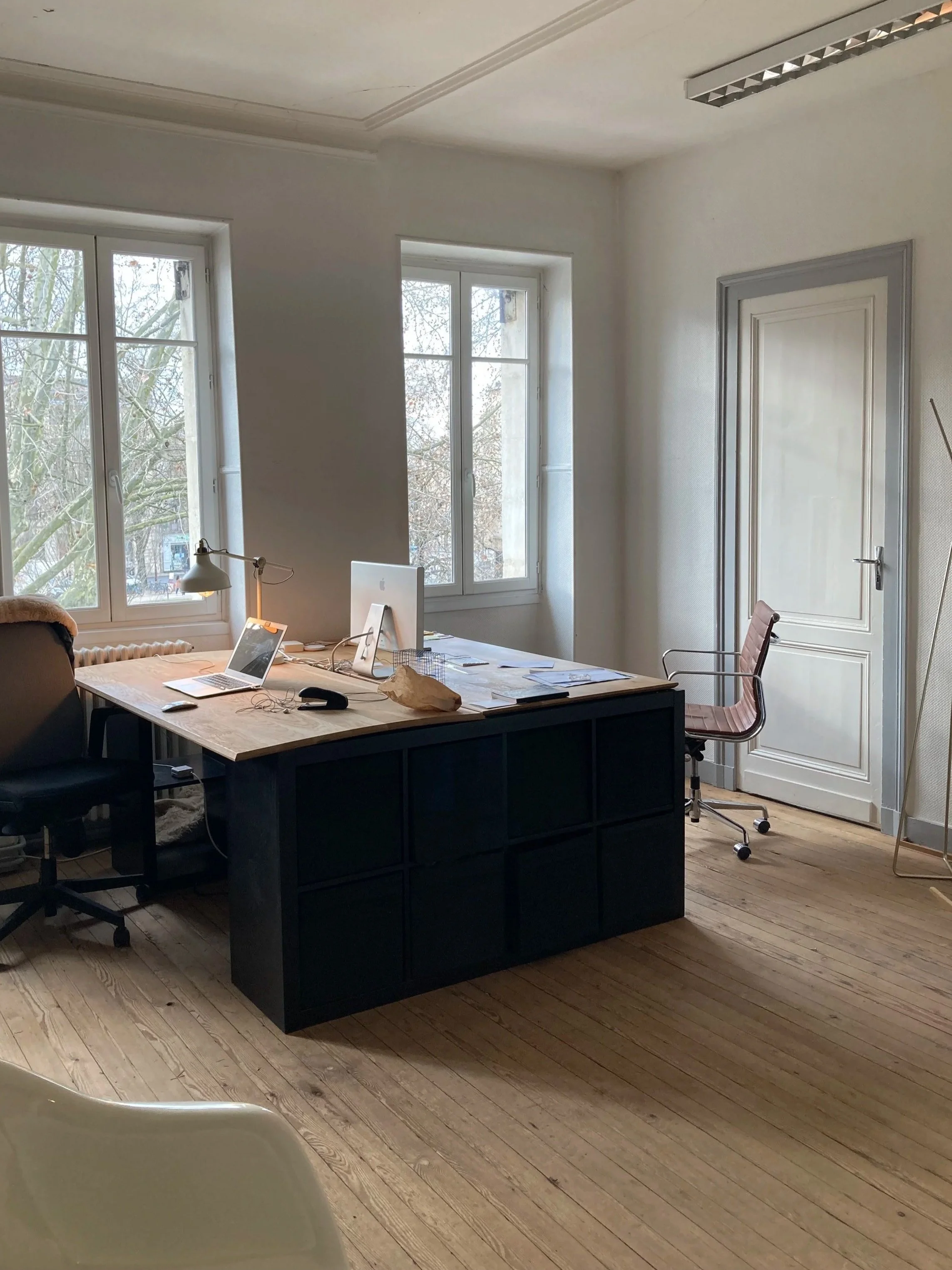 Bureau avec deux fenêtres, un bureau en bois avec un ordinateur, une lampe, un ordinateur portable, des papiers et un sac en papier. Il y a une chaise de bureau en fauteuil autour du bureau. Le sol est en bois clair, la porte est blanche avec des moulures grises, et il y a une chaise blanche partiellement visible en premier plan.