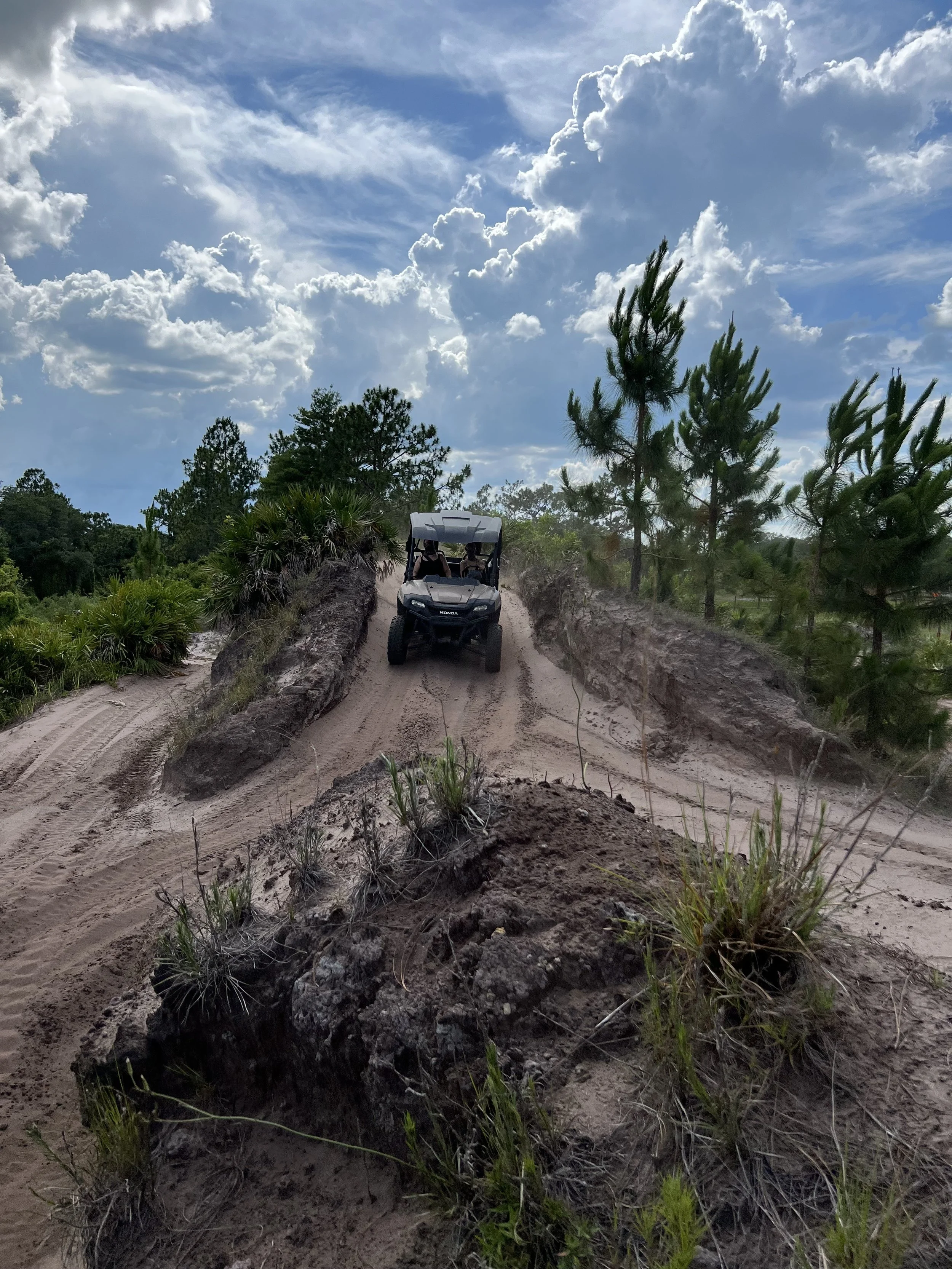 An off-road vehicle driving on a dirt trail surrounded by green trees and plants, with a partly cloudy sky overhead.