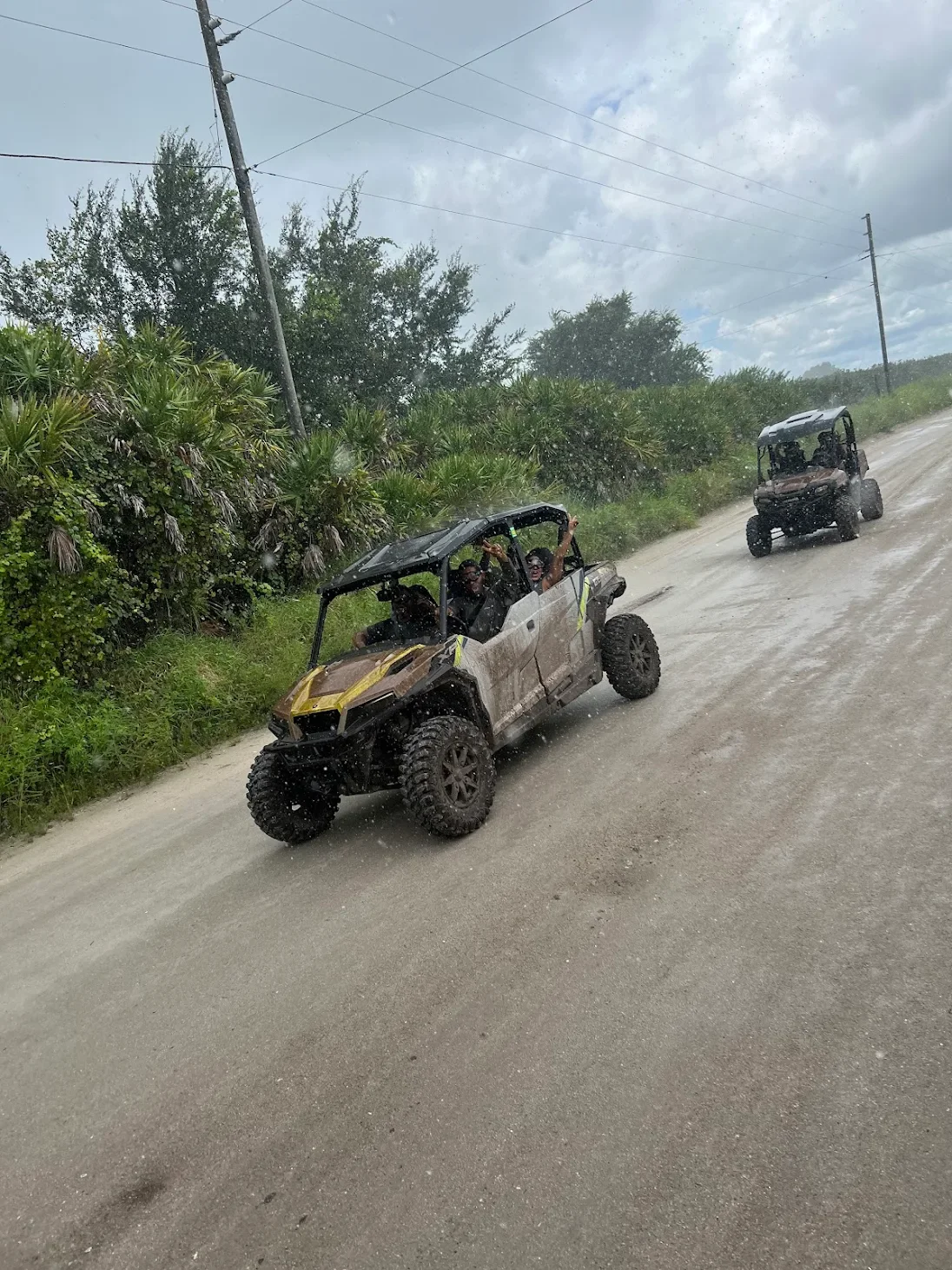 Two people riding in a muddy off-road vehicle on a dirt road, followed by another similar vehicle, with green bushes and cloudy sky in the background, during rainy weather.
