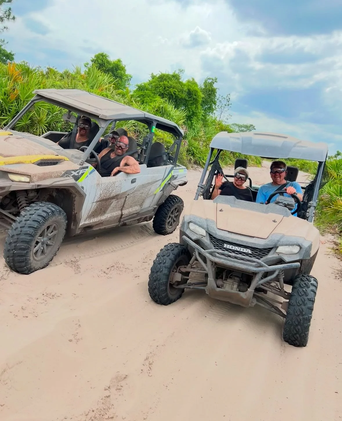 Four people riding in two off-road vehicles on a sandy trail, surrounded by green vegetation and a cloudy sky.