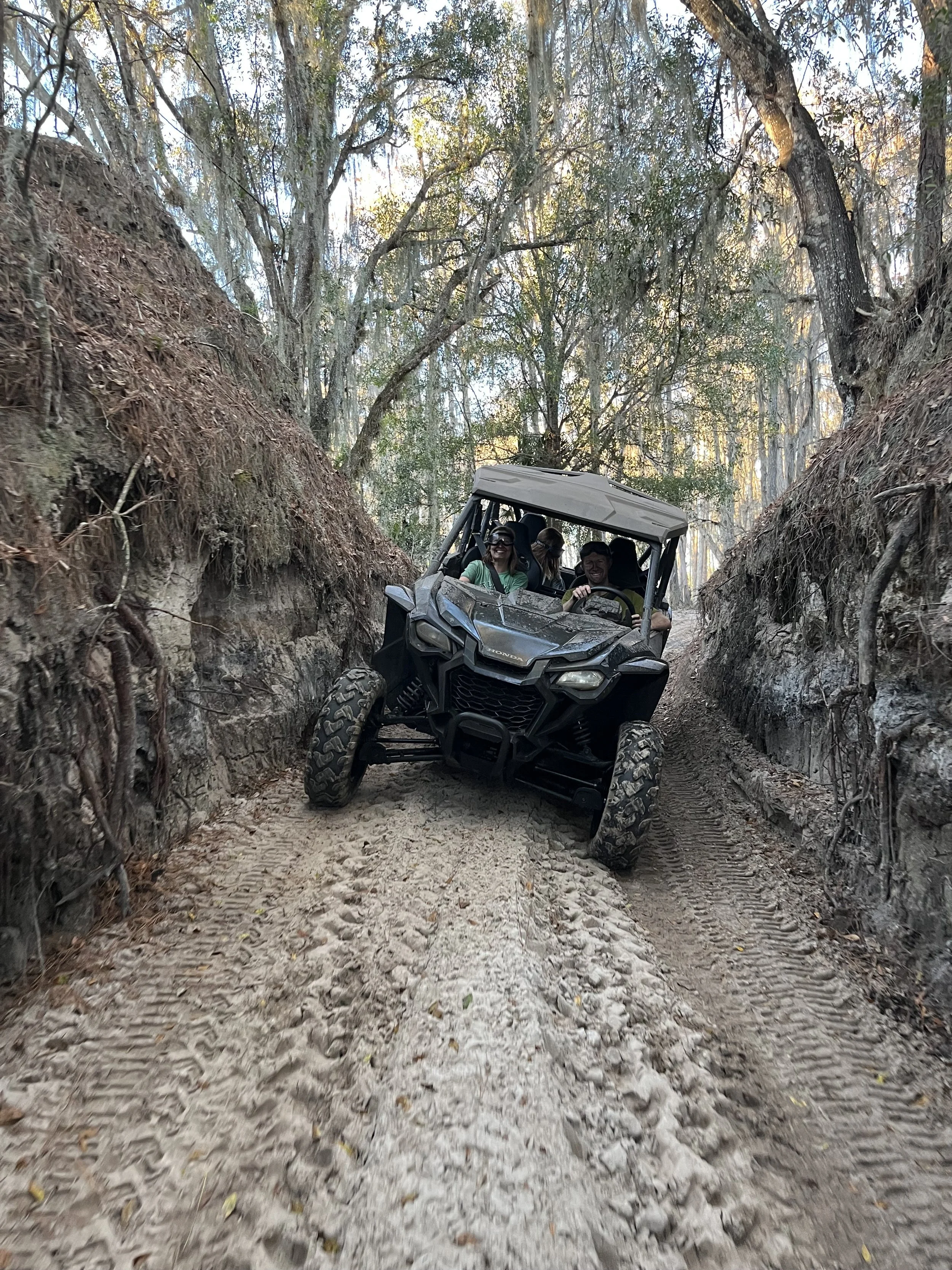 A group of people riding an off-road vehicle through a narrow dirt trail flanked by steep, rocky walls and dense trees.