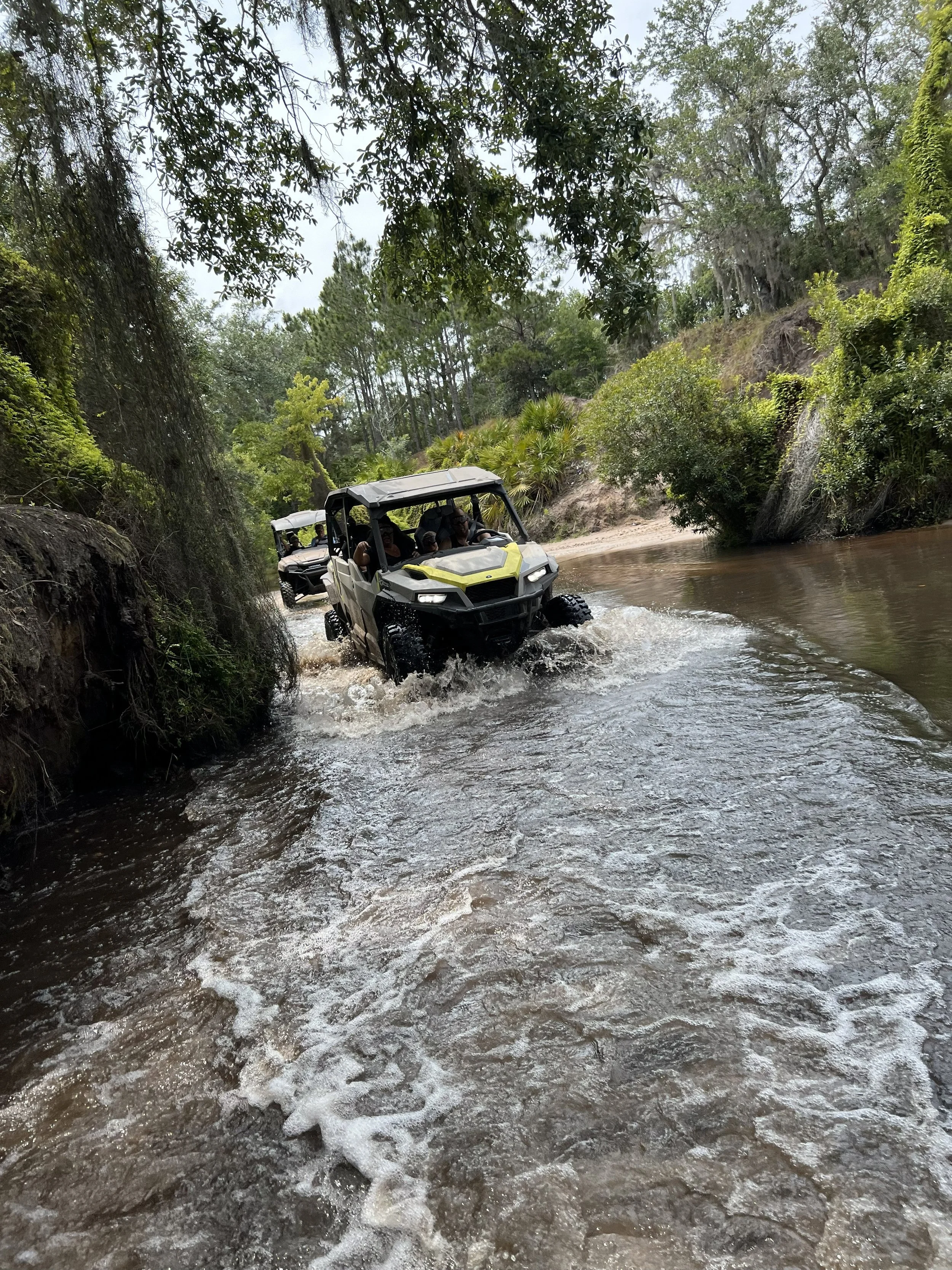 Two off-road utility vehicles driving through a shallow waterstream surrounded by green trees and bushes.