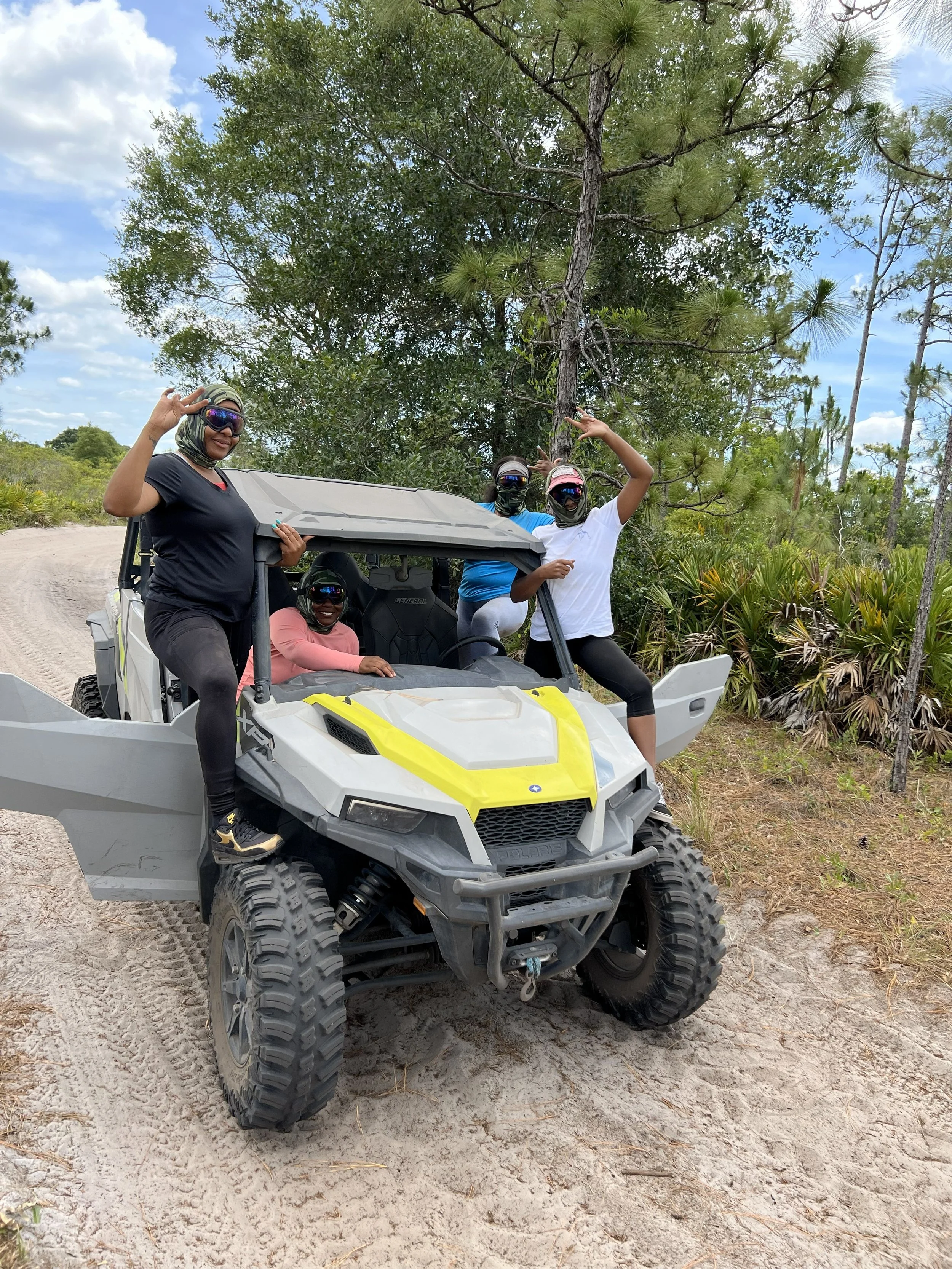 Four women in outdoor gear riding a utility terrain vehicle (UTV) on a dirt trail surrounded by trees and plants, with a partly cloudy sky overhead.