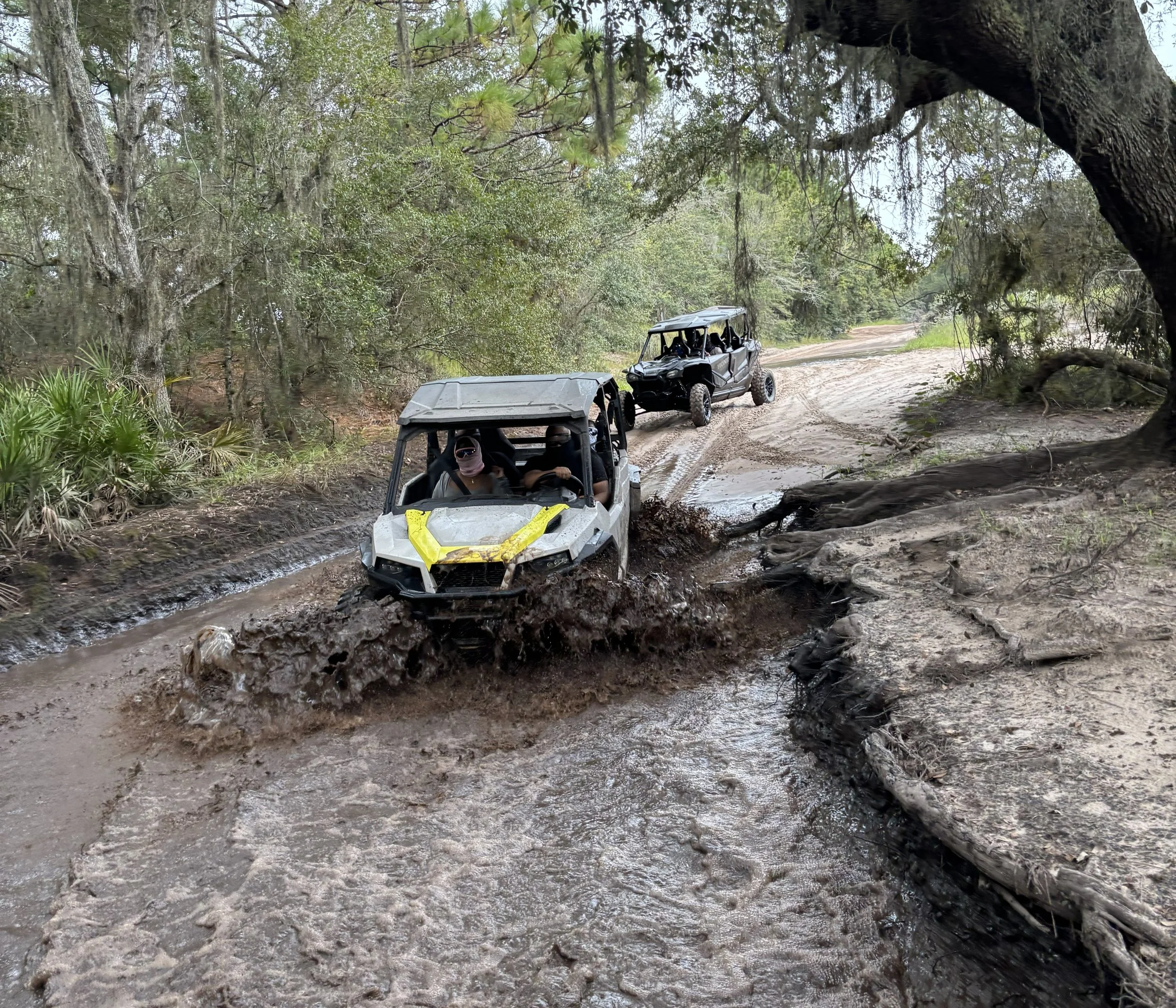 Four off-road vehicles driving through a muddy trail in a forested area.