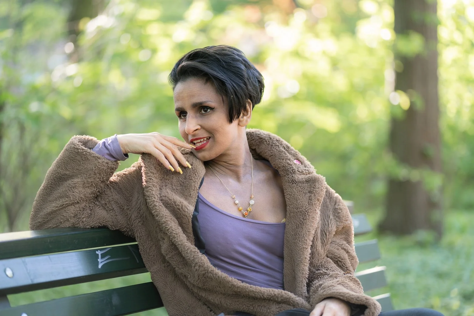 A woman with short black hair wearing a brown fur coat and purple top, sitting on a green park bench with a background of trees and greenery.