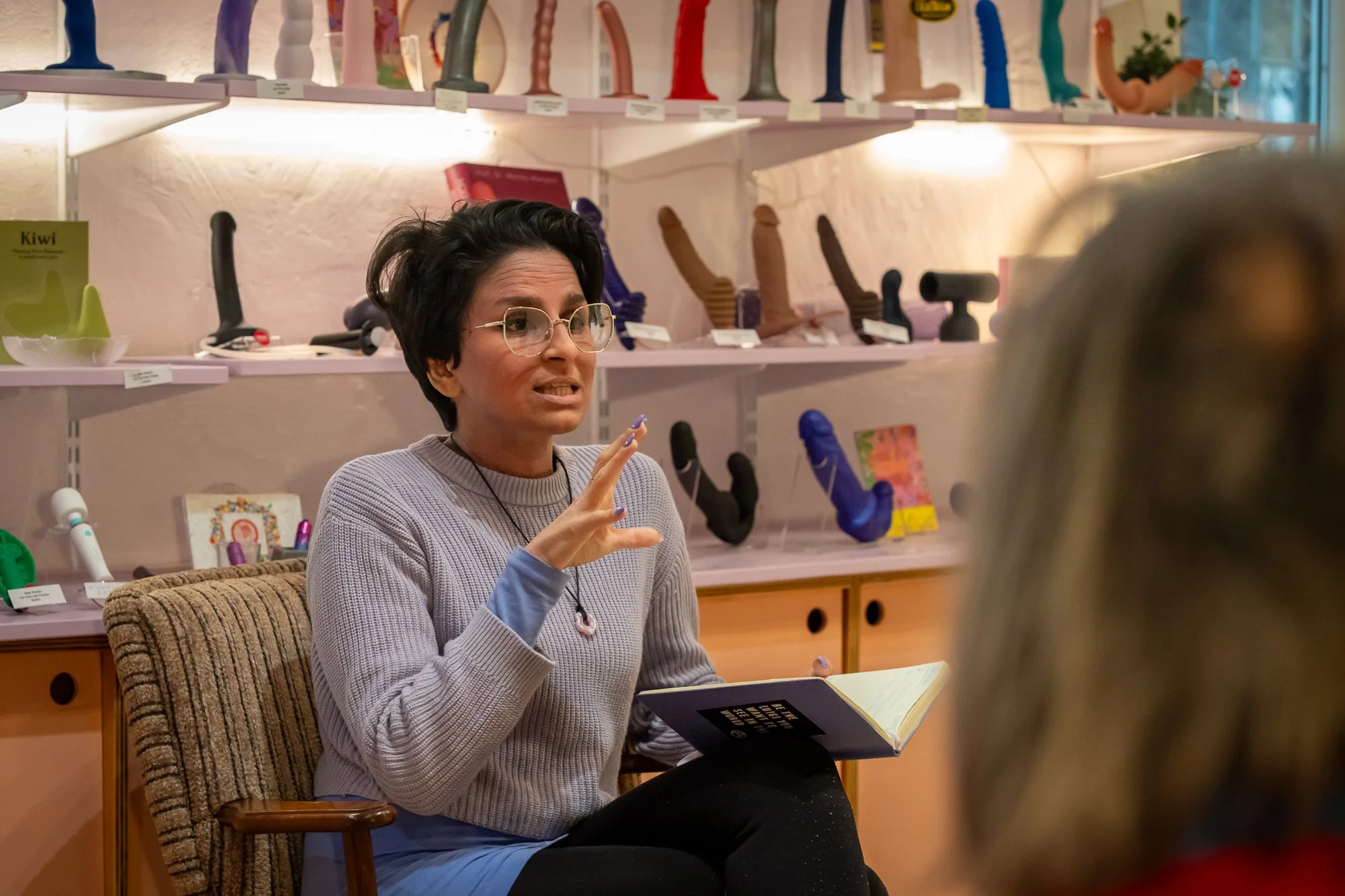A woman with short dark hair, glasses, and a gray sweater gestures while speaking, seated in a chair with a beige cushion. Behind her are pink shelves displaying various colorful silicone dildos and adult toys in store.