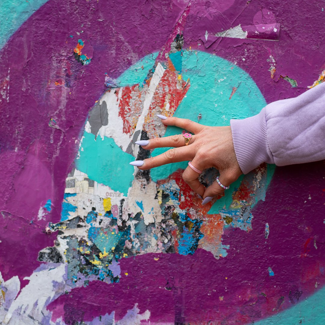 A person with painted nails and rings is touching a worn, colorful street art poster on a purple wall. The poster is torn and peeling, revealing layers of paint and paper underneath.