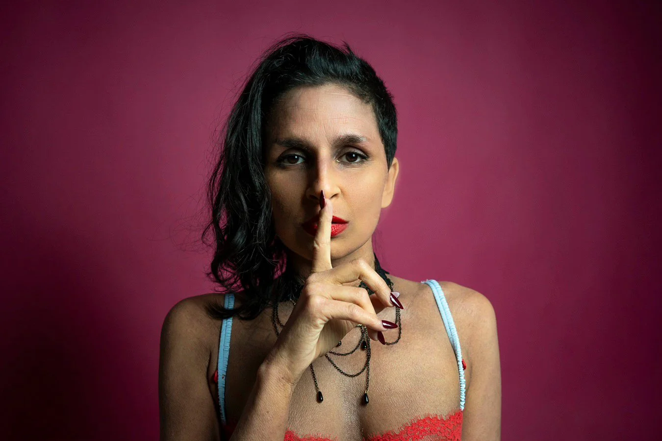 Woman with dark hair in a side style, wearing red lipstick, a black necklace with black beads, and a red top with blue straps, standing against a pink background, holding her index finger to her lips.