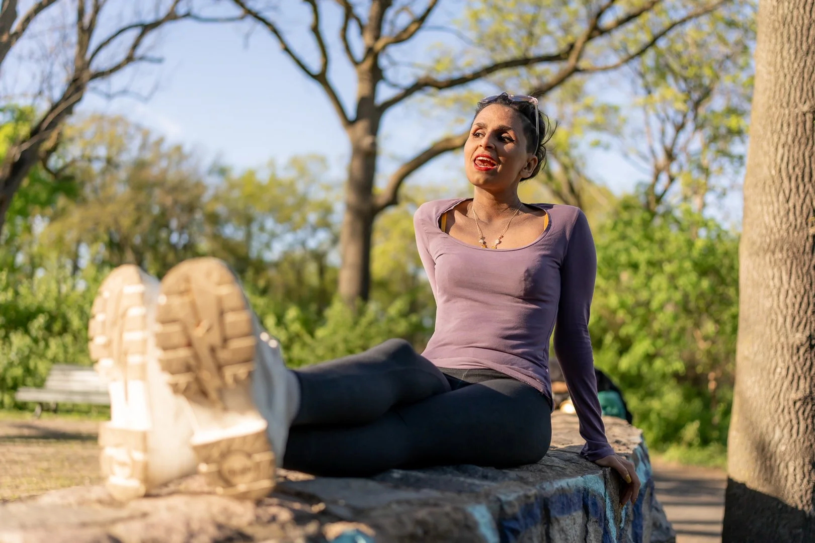 A woman sitting on a stone ledge in a park, wearing a purple long-sleeve shirt, black pants, and sneakers, with her legs extended and crossed at the ankles. She is outdoors with trees and greenery in the background, smiling and chatting.