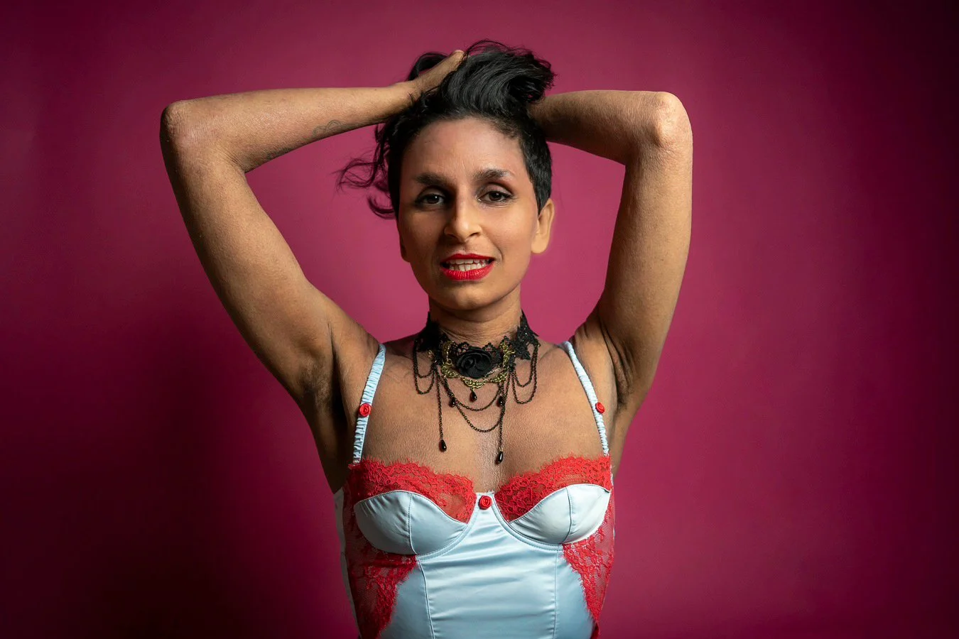 Person with short hair in a lingerie-style top with red and white details, posing with arms raised and hands on head against a pink background, wearing multiple necklaces and red lipstick.