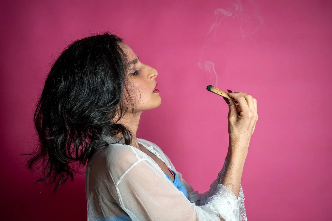 A woman with dark wavy hair and makeup is holding a burning cigarette close to her lips, against a pink background.