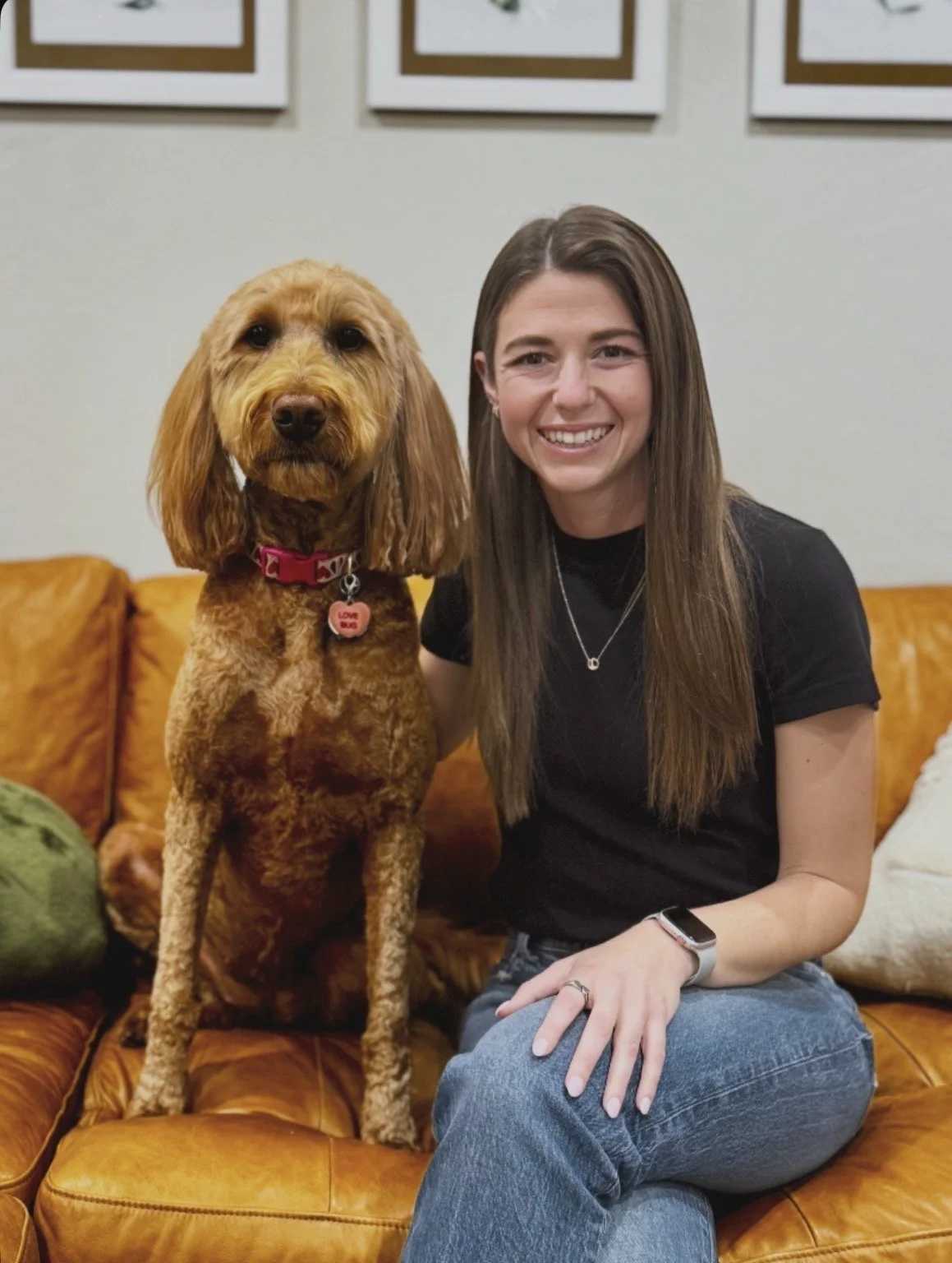 A woman with long brown hair, wearing a black shirt and jeans, sitting on a leather couch next to a brown dog with a pink collar and a bone-shaped tag, in a home setting with framed artwork on the wall. Female mental health counselor and therapy dog.