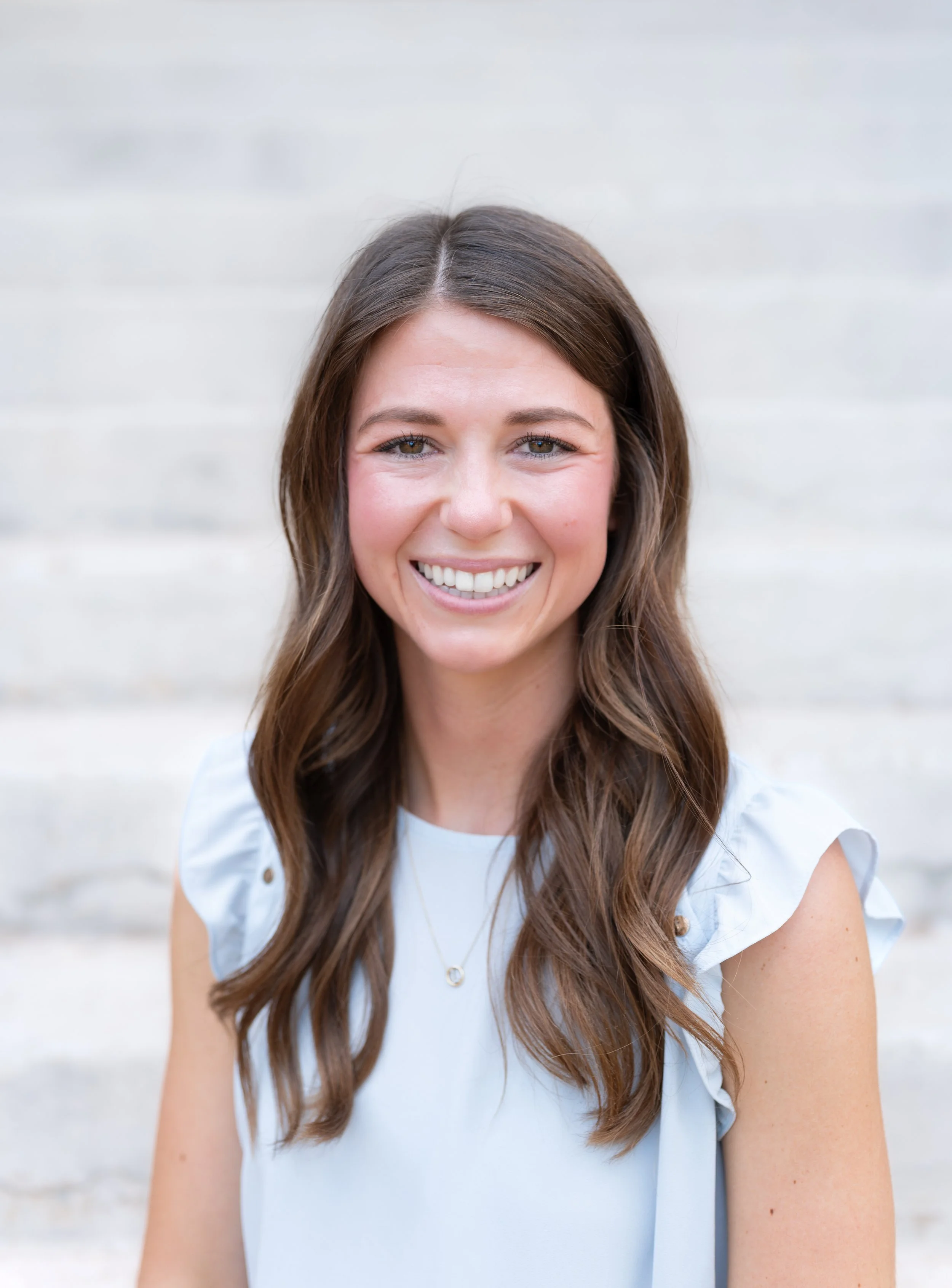 Portrait of a smiling woman with long, wavy brown hair, wearing a white sleeveless top and a delicate necklace, standing in front of a light-colored brick wall.