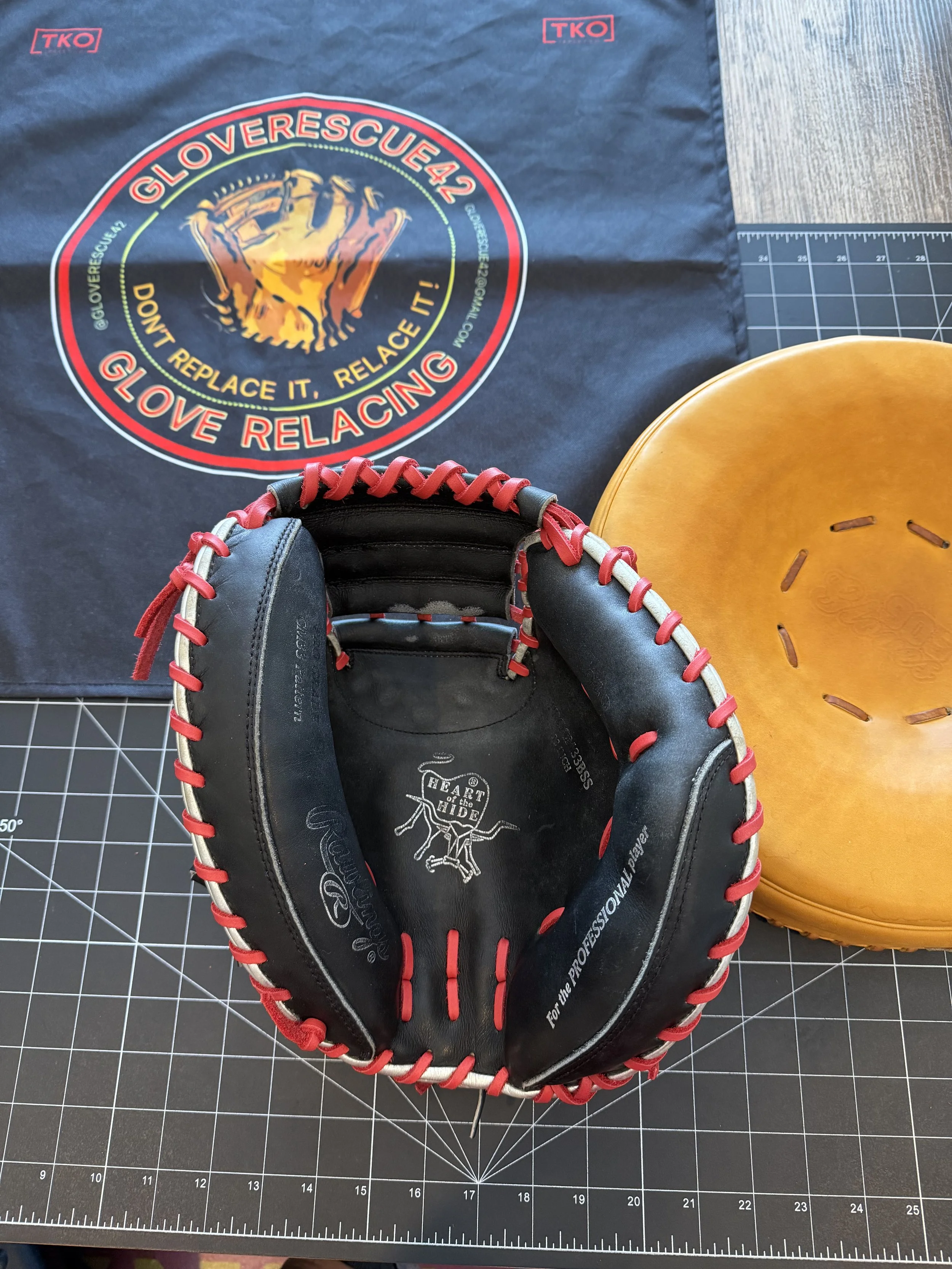 A black baseball catcher's mitt with red and white lacing, placed on a cutting mat. Behind it is a black cloth with a baseball logo, and to the right is a yellowish-brown leather baseball or soft ball.