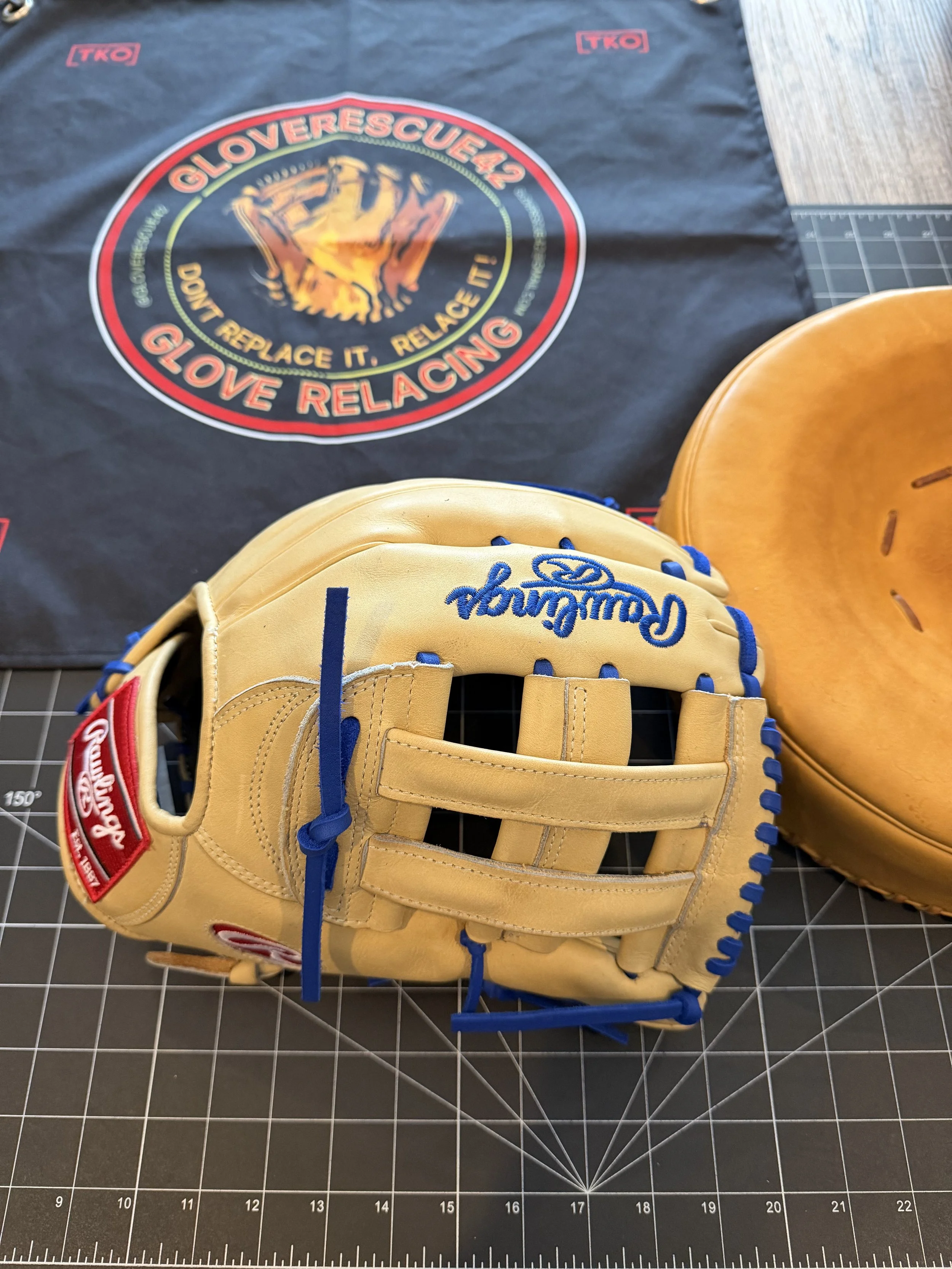 A Rawlings baseball glove, yellow with blue laces, resting on a black cutting mat with measurement grid. Behind the glove is an orange baseball cap and a black cloth with a Glover Rescue 42 logo featuring a raised fist and the text 'Don't replace it. Relace it! Glove relacing'.