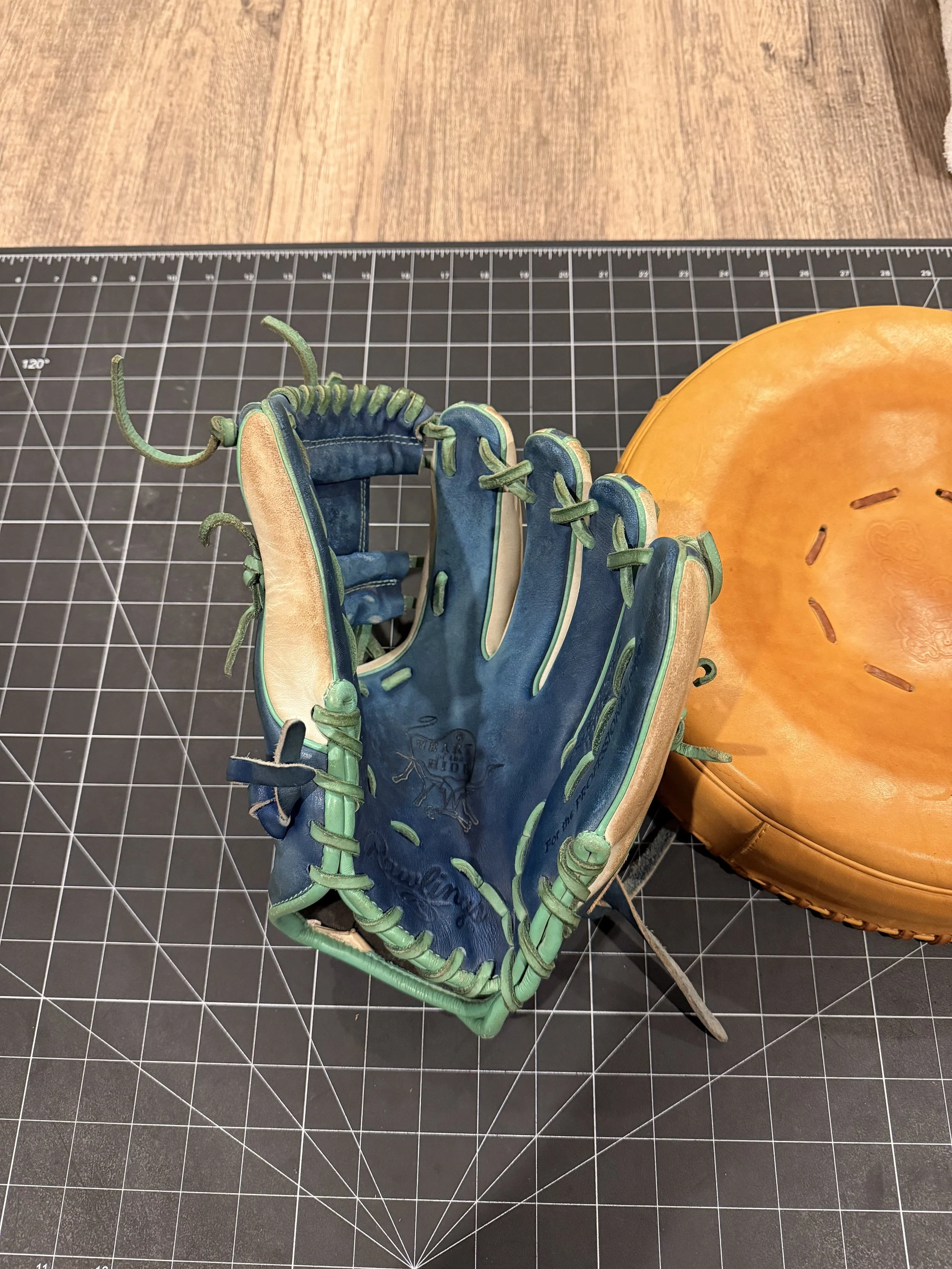 A worn blue and green baseball glove with a beige interior next to a brown baseball. Both are on a black grid cutting mat.
