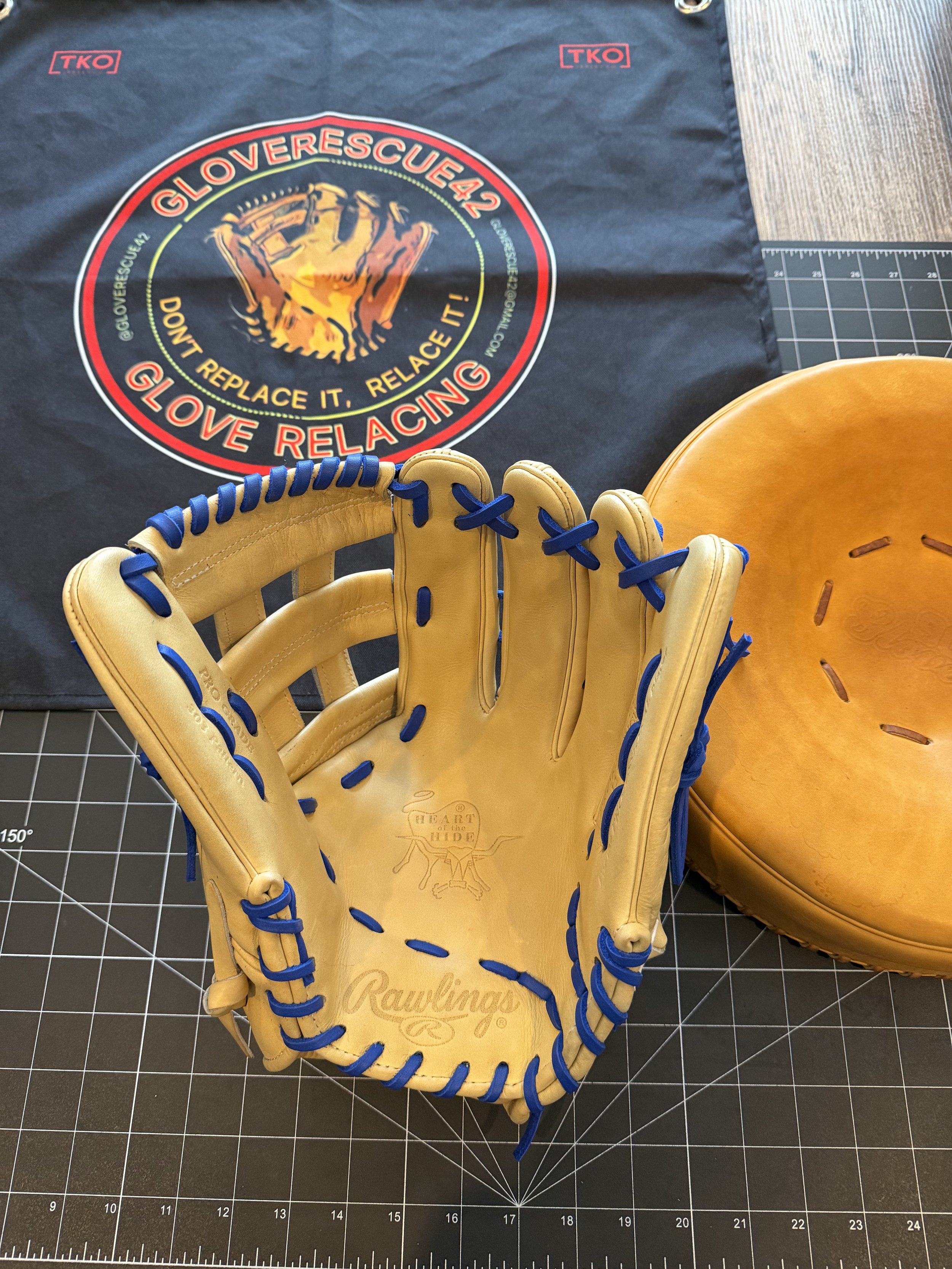 A Rawlings baseball glove, a tan leather catcher's mitt, a black towel with a logo that says 'GLOVERESCUE42, Don't replace it, relace it! GLOVE RELACING,' and a cutting mat on a wooden surface.