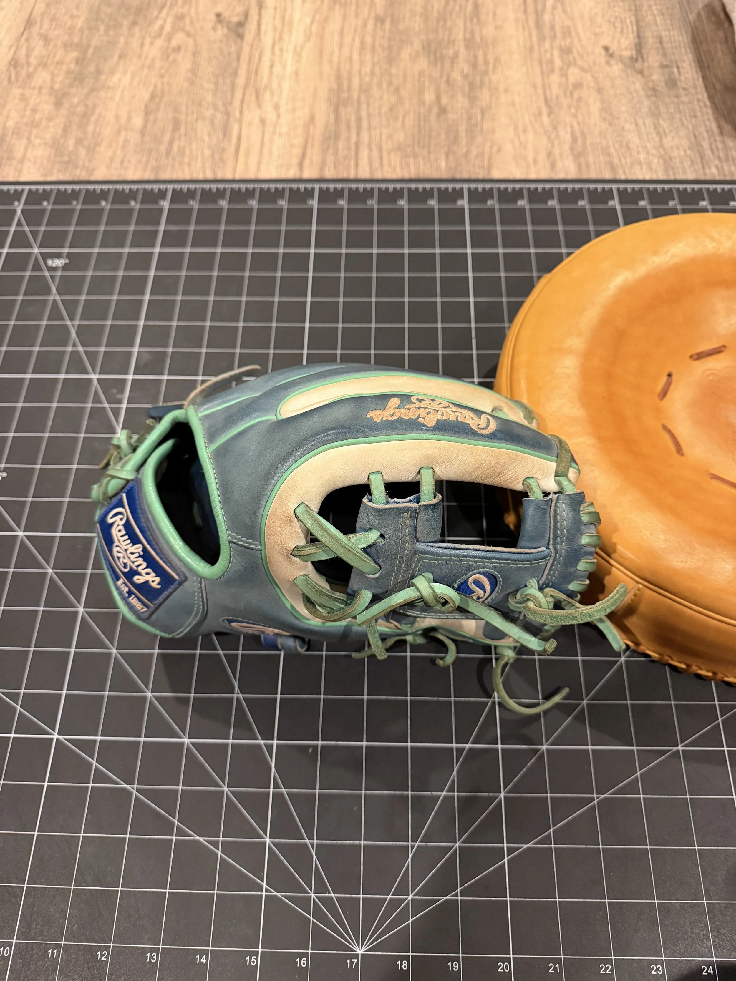 A green and beige baseball glove next to an orange baseball mitt on a black cutting mat with measurement grid.