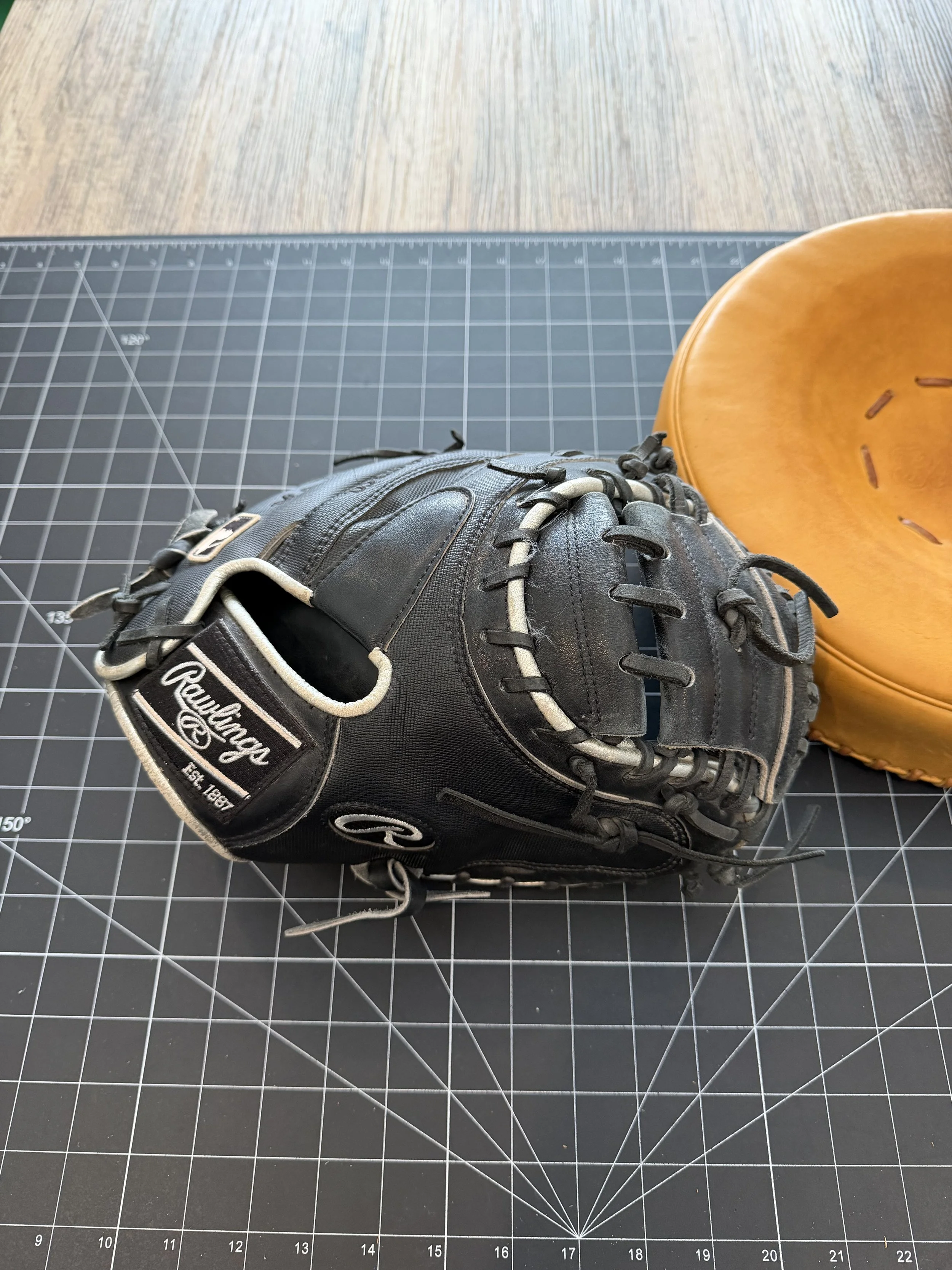 A black Rawlings baseball mitt and a brown baseball glove on a black cutting mat with a grid pattern.