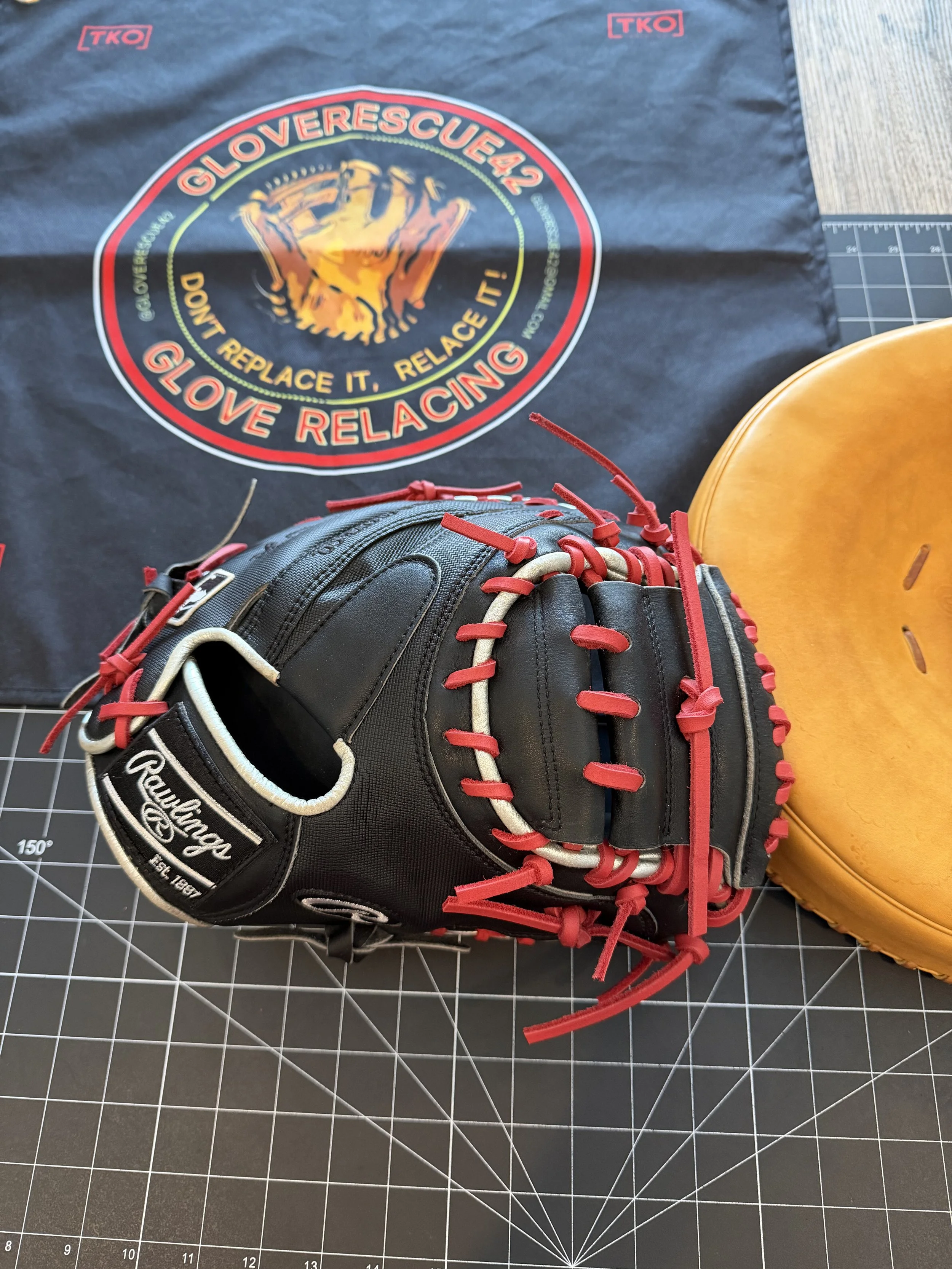 A black Rawlings baseball glove with red laces, placed on a cutting mat, next to an orange baseball mitt. In the background, a cloth with a logo and slogan about glove relacing.