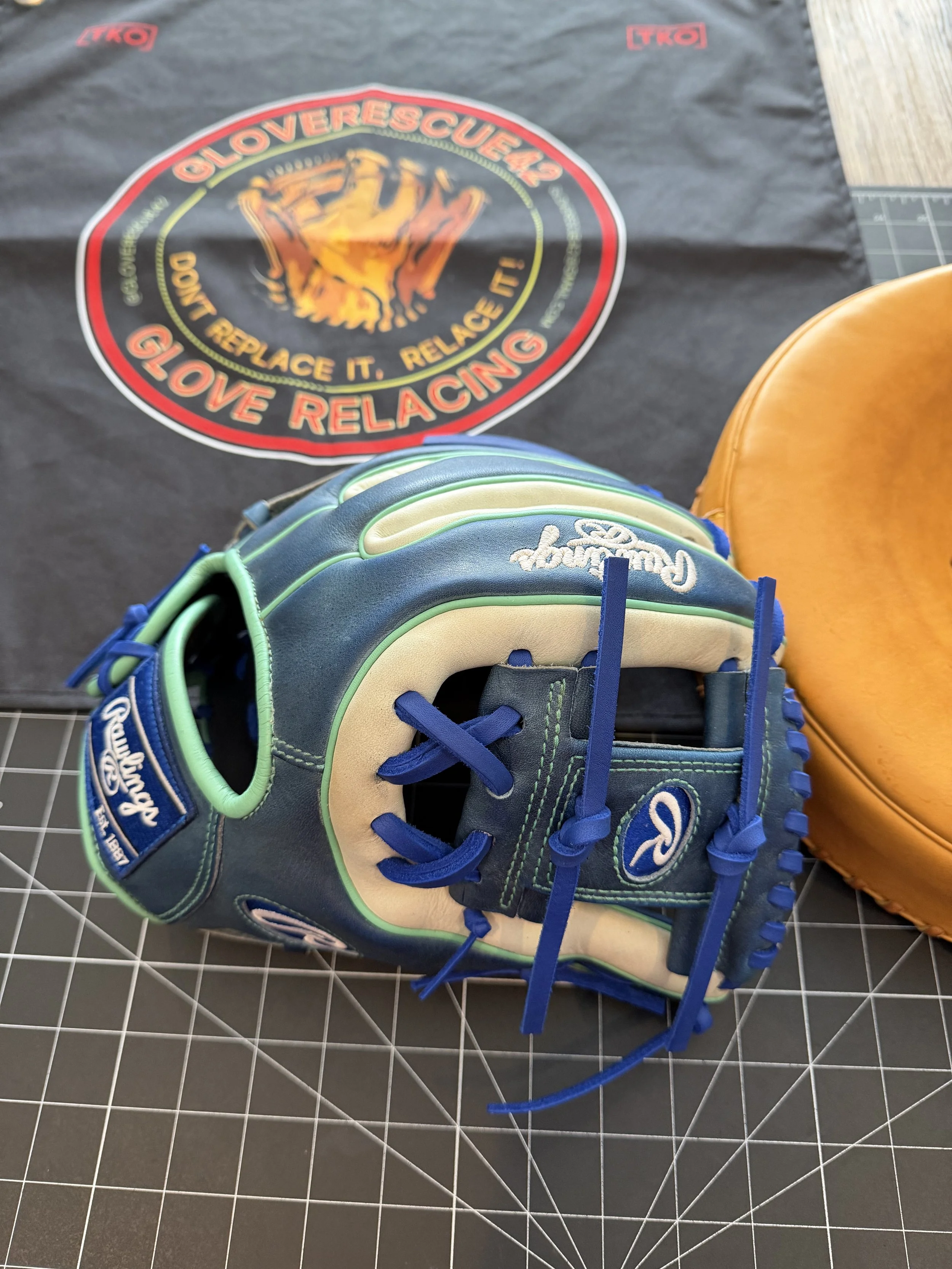 A blue and green baseball glove placed on a grid cutting mat with a black cloth in the background. The cloth features a circular logo with a bear and the words 'Gloverescue24, Don't Replace It. Relace It! GLOVE RELACING'.