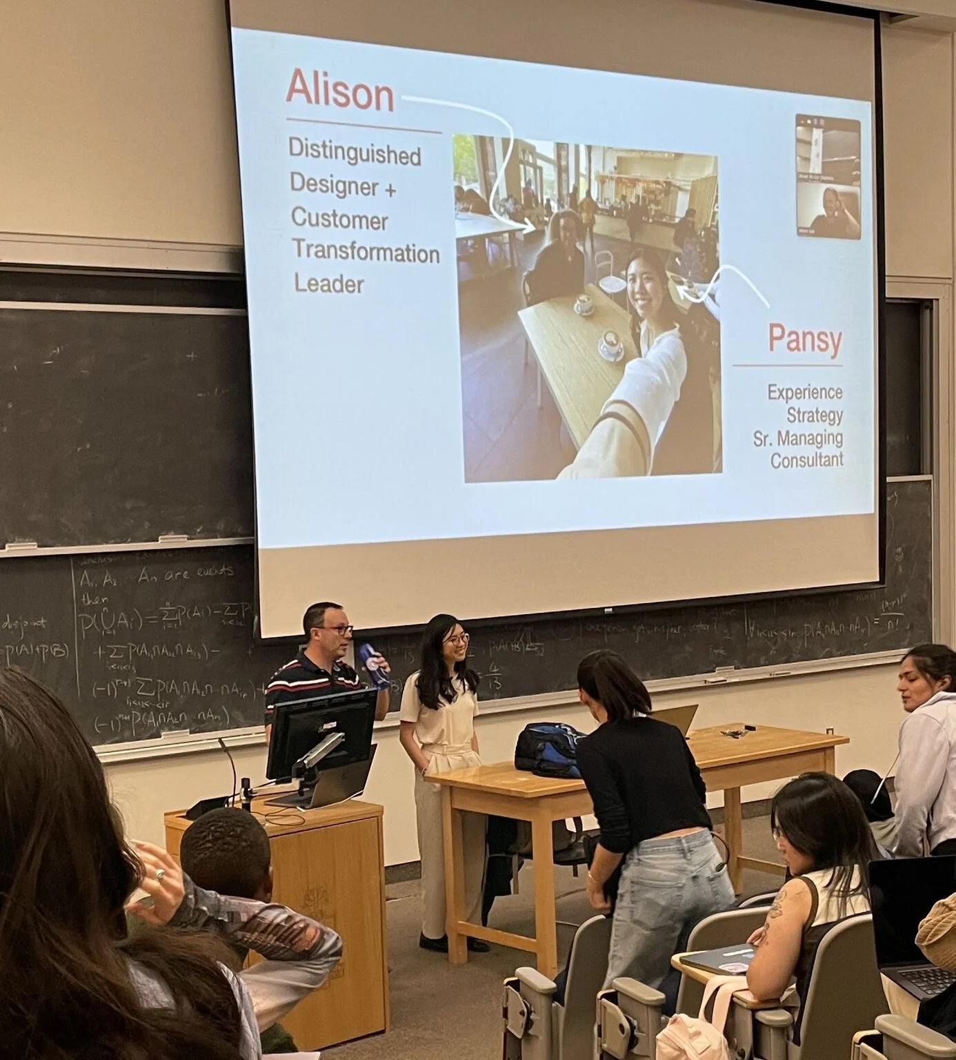 Lecture in a classroom with a large presentation screen showing two women, one labeled Alison, a design leader, and the other Pansy, a senior consulting strategist, taking a selfie. Several students are seated, some taking notes and others using laptops, while a man and a woman stand at the front, preparing to speak or present.