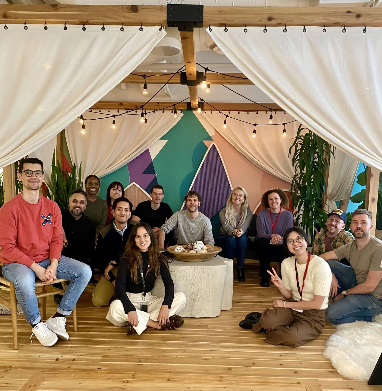 A diverse group of fifteen people gathered indoors, sitting and kneeling on a wooden floor in front of a colorful, abstract wall mural, with white curtains, string lights, and tall green plants in the background at the Pinterest office.